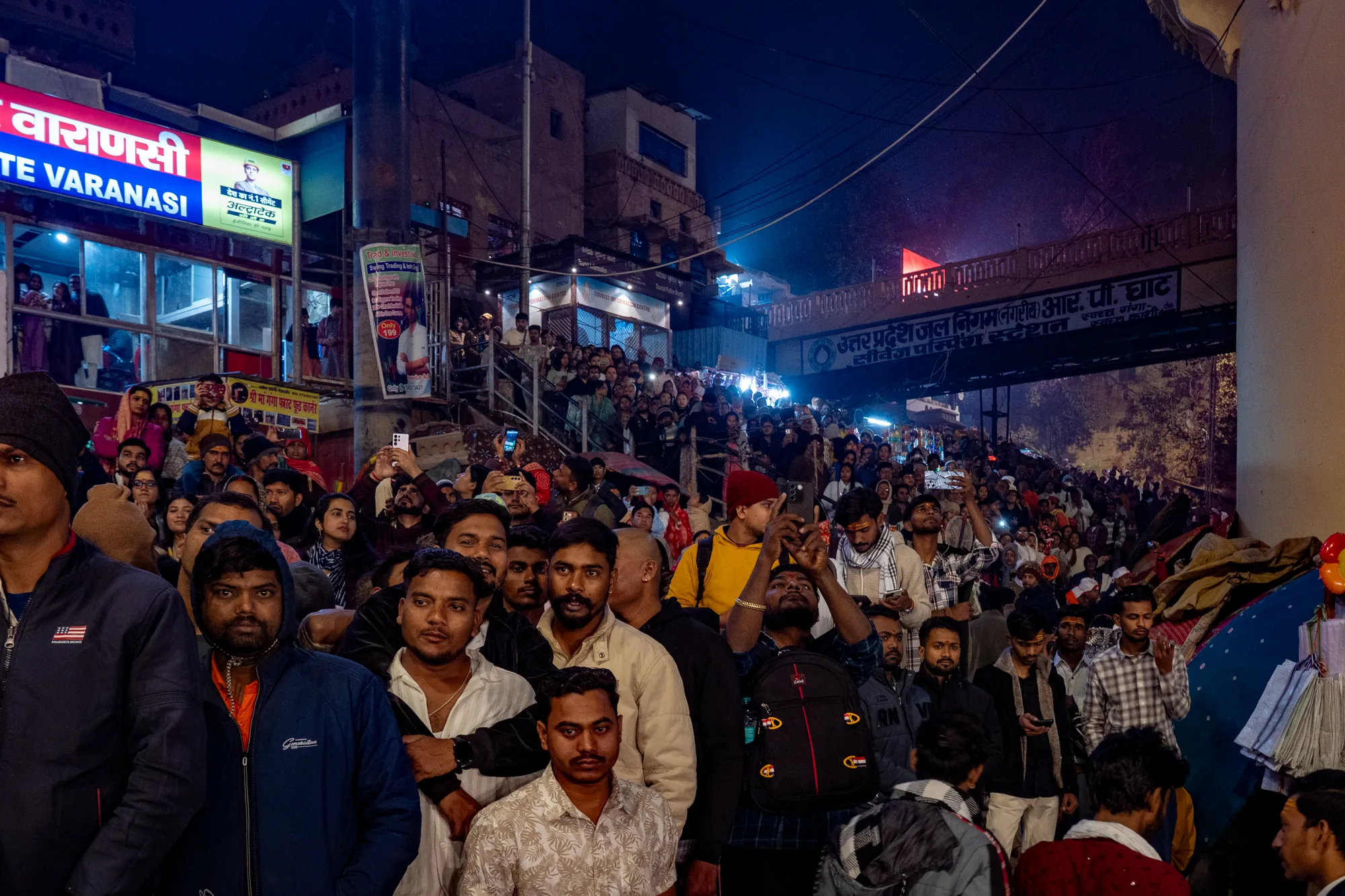 この写真はインドのバラナシで撮影された夜景です。画面全体を埋め尽くすように、非常に多くの人々が集まっています。人々は様々な服装をしており、多くはカメラやスマートフォンを手に何かを見ているようです。建物の看板には「वाराणसी (VARANASI)」と書かれており、観光案内所や飲食店があることがわかります。橋のような構造物には「उत्तर प्रदेश जल निगम नगरीय) आर. पी. घाट」と書かれた看板があり、上下水道に関する施設のようです。夜の暗さと建物の明かり、そして人々の熱気が混ざり合い、バラナシ独特の賑わいを伝えています。
