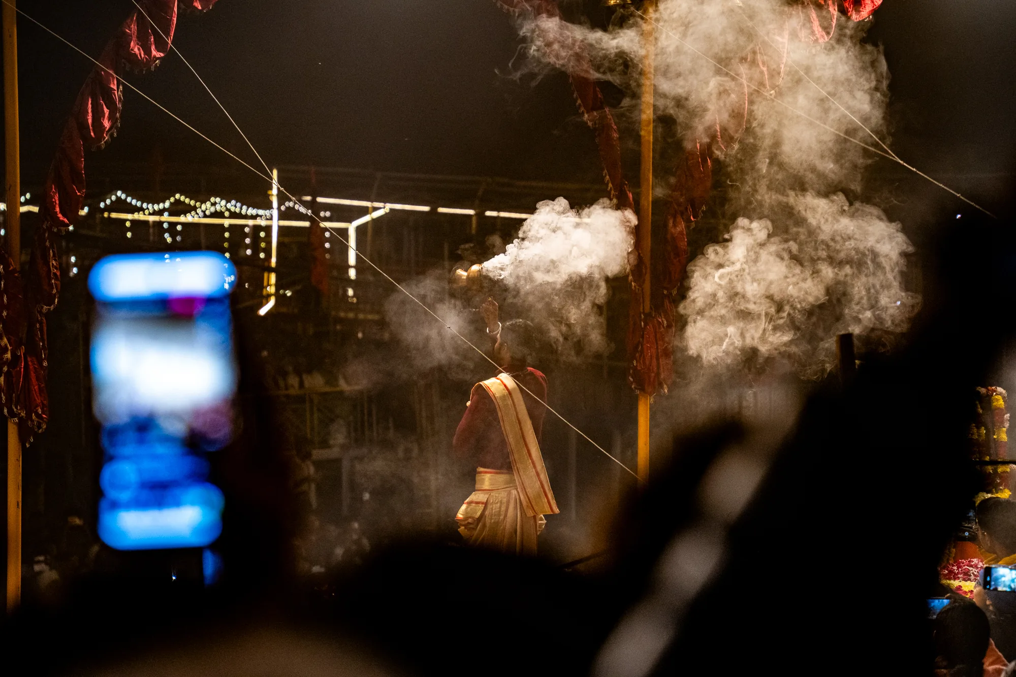 この写真は、インドのバラナシで撮影された夜の宗教儀式の様子を捉えています。中央には、伝統的な衣装を身に着けた人物が、香炉のようなものを持って立っており、そこから白い煙が立ち上っています。煙は周囲に広がり、神秘的な雰囲気を醸し出しています。背後には、赤い布が飾られた柱や、ぼんやりとした光を放つ装飾が見えます。写真の手前には、人々のシルエットが写っており、多くの人がスマートフォンで写真を撮っている様子が伺えます。暗い背景と立ち込める煙によって、厳かで幻想的な雰囲気が強調されています。