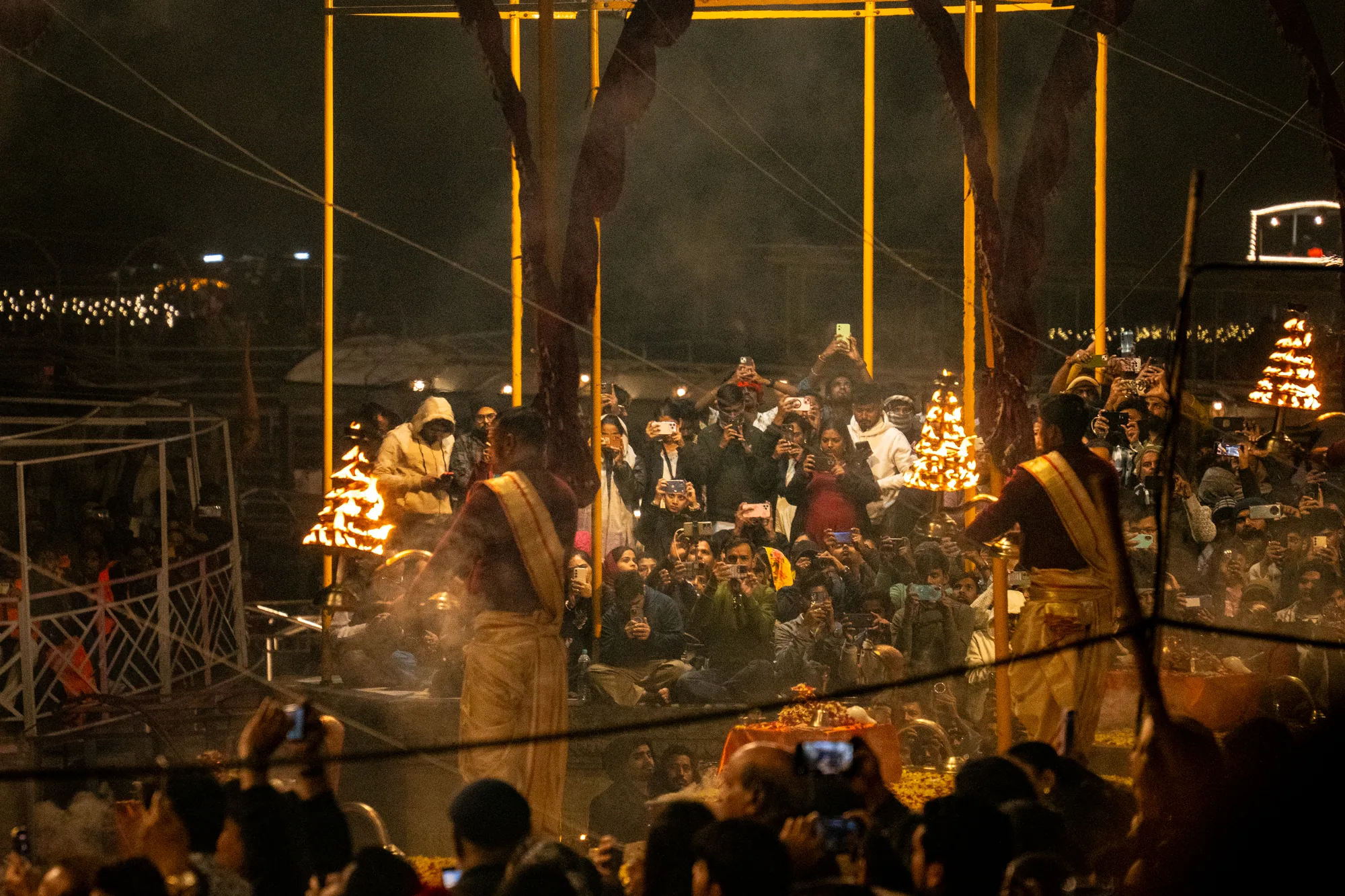 この写真はインドのバラナシで撮影された夜の風景です。画面全体に大勢の人が密集しており、多くの人がスマートフォンやカメラを構え、何かの儀式を撮影しているようです。写真の中央には、伝統的な衣装を身につけた人物が二人おり、それぞれが燃え盛る松明のようなものを持っています。背景には、黄色い光を放つ金属製の柱が立っており、煙が立ち込めています。周囲には、ガート（沐浴場）のような構造物も見えます。全体的に、神秘的で熱気に満ちた雰囲気が漂っており、異文化の儀式を見物する人々の興奮が伝わってくるようです。観客は、若い世代から年配の世代まで、さまざまな人がいるように見えます。