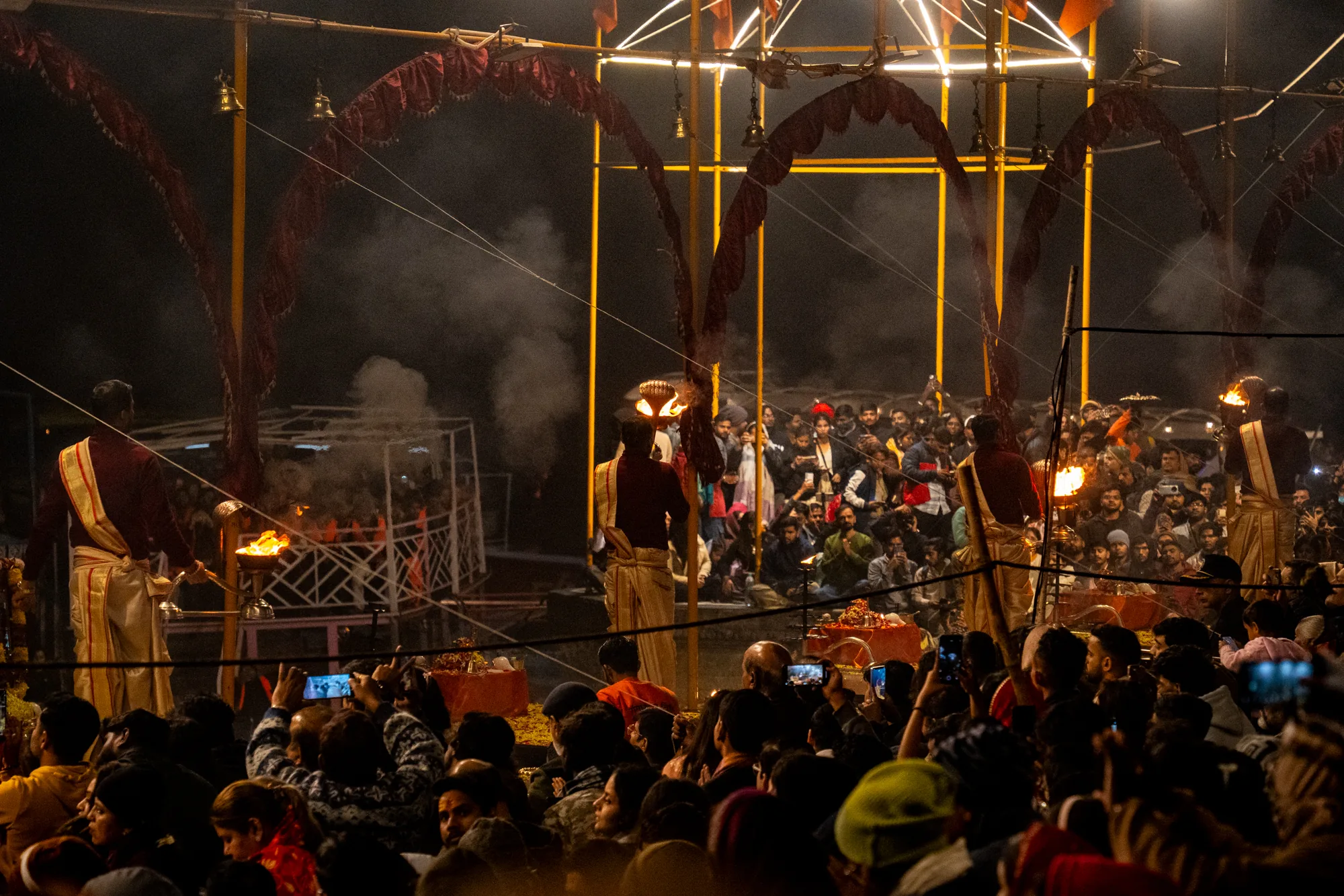 この写真はインドのバラナシで撮影されたもので、夜に行われている儀式の様子を捉えています。写真全体は暗い色調で、周囲の暗闇と儀式で使用されている炎の光が印象的です。  手前には大勢の観客がおり、人々は儀式の様子をスマートフォンなどで撮影しています。観客の頭越しに見えるのは、儀式を執り行う人々の姿です。彼らは伝統的な衣装を身に着け、燃え盛る炎を持っています。炎は暗闇の中で明るく輝き、煙が立ち上っています。  背景には、装飾が施された金属製の構造物が見えます。構造物はライトアップされており、その光が周囲を照らしています。構造物の周りにも人々が集まっており、儀式の様子を見守っているようです。写真全体から、神聖で神秘的な雰囲気が伝わってきます。人々の熱気と、炎の力強さが感じられる一枚です。