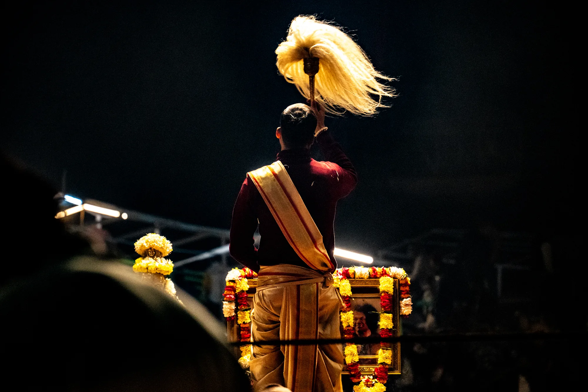 インドのバラナシで撮影された写真です。暗闇の中で、祭壇の前で儀式を行っている男性の後ろ姿が写っています。男性は赤いシャツに金の縁取りがある白い腰布を身に着けており、同じく金の縁取りがあるストールを肩にかけています。頭の上には、光を浴びて輝く、大きな白い毛束のようなものを持っています。祭壇は金色の額縁で飾られており、マリーゴールドのような花で飾られています。額縁の中には人物の写真が見えます。全体的に厳かで神秘的な雰囲気が漂っています。
