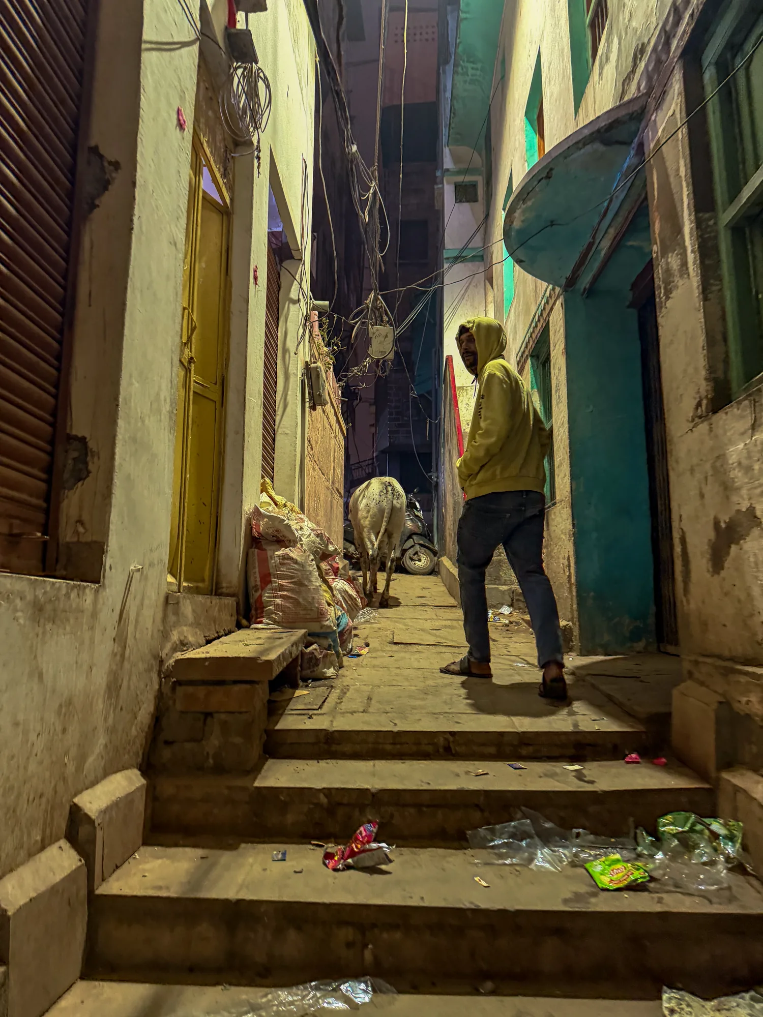 写真はインドのバラナシの夜の路地裏を捉えています。路地は狭く、左右には古い建物が並び、複雑に絡み合った電線が上空を覆っています。路面の階段にはゴミが散乱しています。路地の中央には牛が一頭立っており、その手前にはフード付きの服を着た男性がカメラの方を振り返っています。男性はジーンズとサンダルを着用しており、リラックスした様子です。路地の奥にはバイクが停まっているのが見えます。写真全体は黄昏時の人工的な光に照らされ、やや雑然としていますが、同時に独特な活気を感じさせます。建物の壁は古く、所々に補修の跡が見られます。階段状の路面や建物の色合いから、この場所の歴史と文化が感じられます。