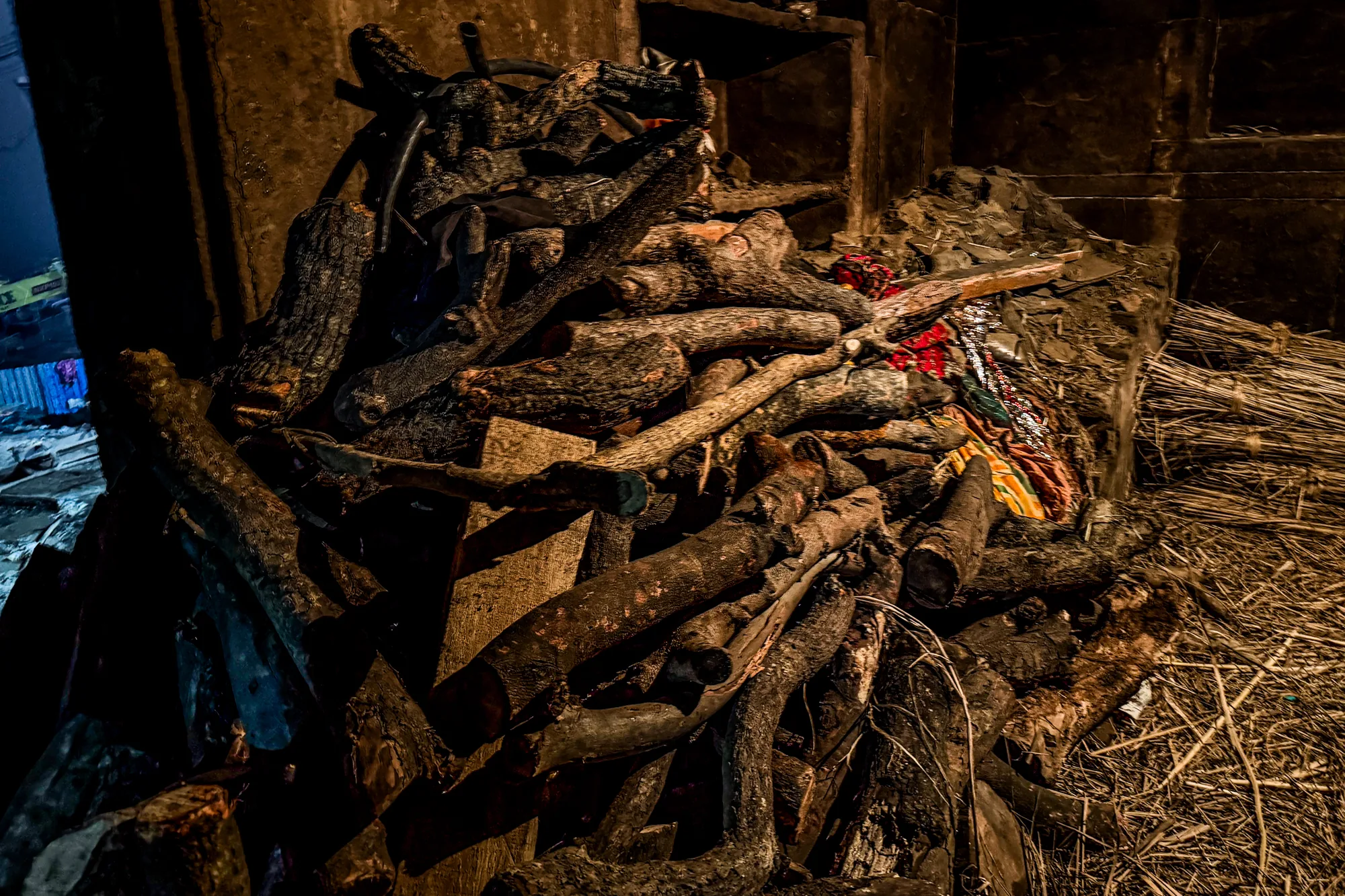 この写真は、インドのバラナシで撮影されたもので、おそらく火葬に使われる薪の山を捉えています。画面全体に、様々な太さ、長さの薪が積み上げられており、その多くは表面が焦げ付いたように黒ずんでいます。薪の質感は、荒々しく、使い込まれた印象を与えます。薪の山の上には、布のようなものが見え、鮮やかな色彩が僅かに確認できます。また、薪の山の右側には、乾燥した草のようなものが束ねて積み上げられており、全体的に埃っぽく、乾燥した雰囲気です。周囲の環境は暗く、ほとんど光がありません。石造りの壁の一部が見え、背景に溶け込むように暗くなっています。全体的に、死や宗教的な儀式を連想させる、厳粛で重々しい雰囲気の写真です。