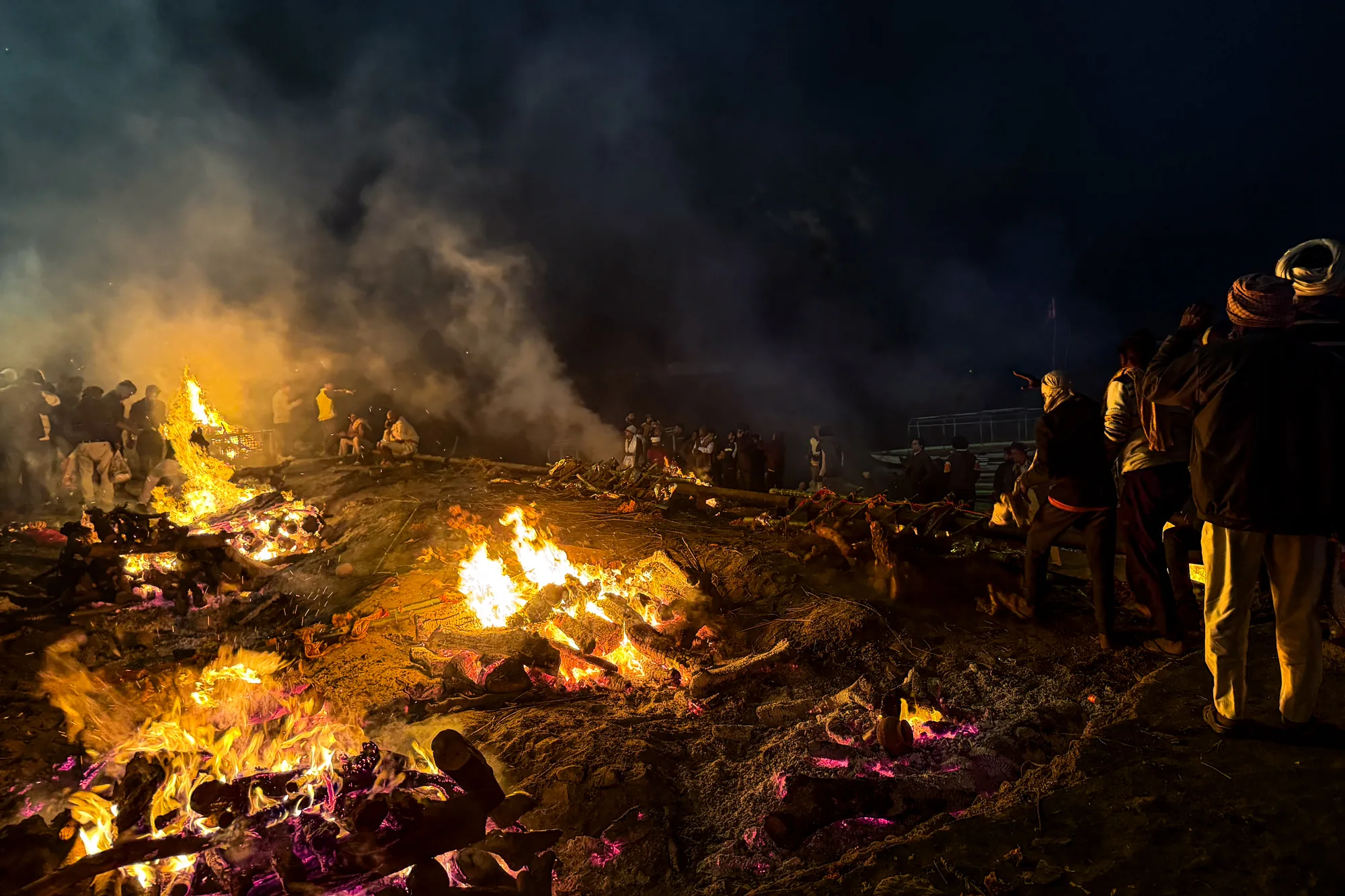 インドのバラナシで撮影された写真です。夜の暗闇の中、燃え盛る炎が印象的です。複数の薪が組まれ、大きな炎が立ち上がっています。炎の周りには人々が集まっており、火葬の様子を見守っているようです。煙が空に立ち上り、全体的に厳かな雰囲気を醸し出しています。画面右側には、炎を見つめる人々のシルエットが確認できます。地面は暗く、燃え残った薪や灰が散らばっているのが見えます。全体的に暗い色調ですが、炎のオレンジ色や黄色が際立ち、強いコントラストを生み出しています。これは、バラナシのガンジス川沿いにある火葬場の様子を捉えた写真と思われます。