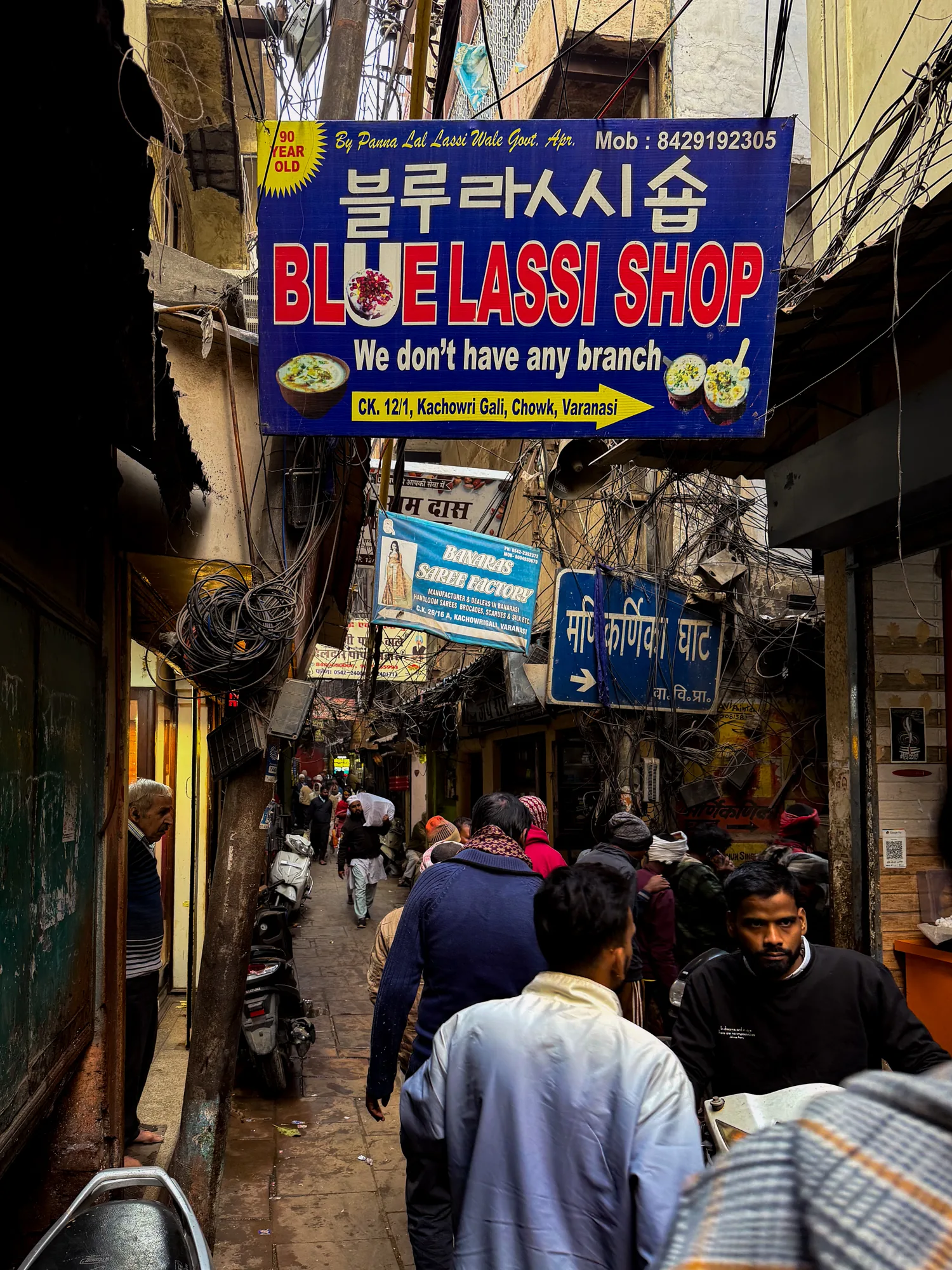 この写真はインドのヴァラナシで撮影された、賑やかな路地裏の様子を捉えています。狭い路地は多くの人々で混み合っており、生活感にあふれた雰囲気です。写真手前には人々が歩いており、その服装は様々です。奥にはさらに多くの人が行き交い、混沌とした活気が感じられます。  路地の両側には建物が立ち並び、壁には看板やポスターが所狭しと貼られています。特に目を引くのは、韓国語も併記された「BLUE LASSI SHOP」というラッシー店の看板です。また、電線が複雑に絡み合い、空を覆うように張り巡らされており、インドらしい雑多な風景を形作っています。スクーターが数台、路地の脇に停められており、人々の生活の足となっていることが伺えます。路地裏の薄暗さと、人々の熱気が混ざり合い、独特な雰囲気を醸し出している写真です。