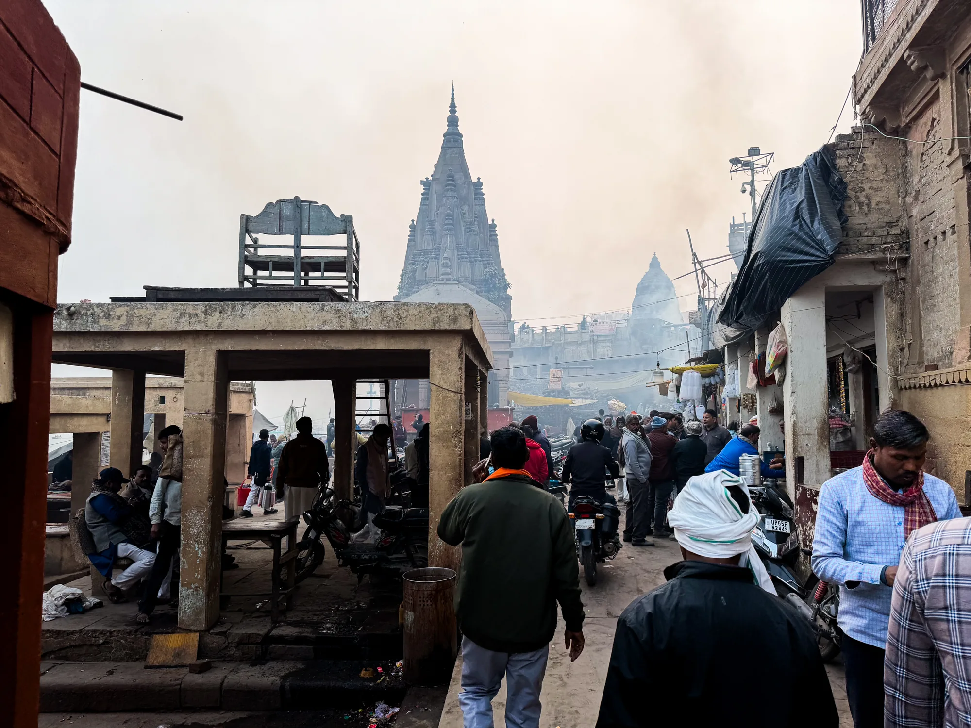 写真はインドのバラナシで撮影されたもので、狭い通りが人やバイクで賑わっている様子が捉えられています。通りの奥には、巨大な寺院の尖塔がそびえ立っており、独特の景観を作り出しています。空は薄い灰色で、煙のようなものが漂っており、全体的に霞んだ雰囲気を醸し出しています。通りの両側には古い建物が立ち並び、壁は剥がれかけ、ところどころに補修の跡が見られます。写真手前には、柱で支えられた建物の一部があり、人々が座って休憩している様子が見られます。通りの人々は様々な服装をしており、インドの日常的な風景を切り取っているようです。全体的に、生活感と宗教的な雰囲気が混ざり合った、バラナシの独特な雰囲気が感じられる写真です。
