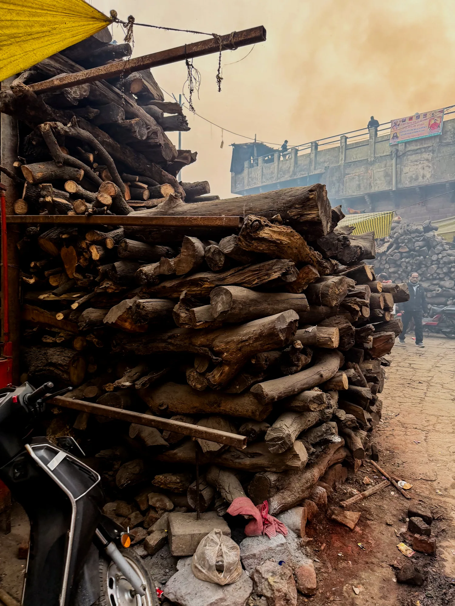 インドのバラナシで撮影された写真です。画面の大部分を占めるのは、大量に積み上げられた薪です。薪は様々な太さの木材で構成されており、乾燥した茶色をしています。左手には、薪の山に寄り添うように黒いスクーターが置かれています。地面は土と石畳が混ざったような状態であり、ゴミや瓦礫が散見されます。背景には建物が見え、屋上に人がいるのがわかります。空は煙霧に覆われているように見え、全体的に薄暗い雰囲気です。写真全体から、生活感と独特の雰囲気が感じられます。
