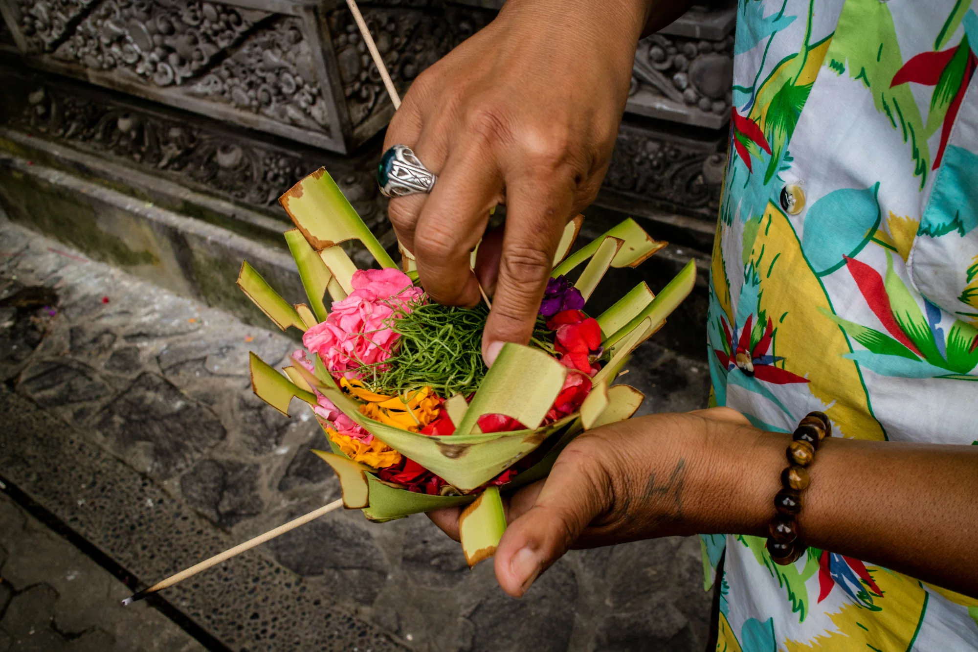 写真はインドネシアのバリ島で撮影されたもので、伝統的なお供え物（チャナン）を準備している様子を捉えています。手元に焦点が当てられており、人物はカラフルなシャツを着ています。両手でお供え物を持ち、片方の手で丁寧に花を配置しているようです。お供え物は、ヤシの葉で作られた器の中に、ピンク、オレンジ、赤、紫の花、そして緑の草のようなものが詰められています。お供え物にはお香も添えられているようです。指には大きな指輪がはめられ、腕にはブレスレットが付けられています。背景には石造りの壁が見え、寺院のような場所で行われていることが伺えます。全体的に、伝統的な儀式や信仰が息づくバリの文化的な雰囲気が伝わってきます。