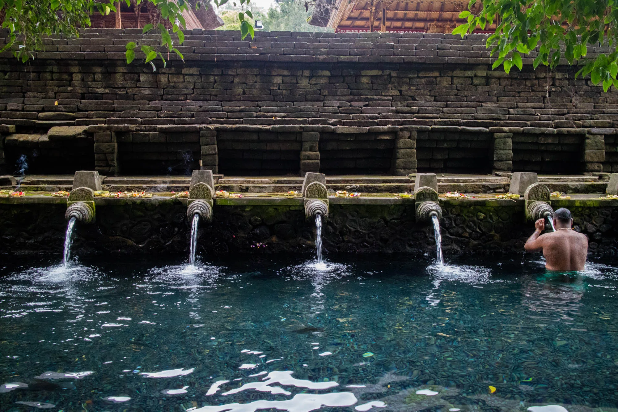 インドネシアのバリ島で撮影された写真です。写真には、古代の石造りの壁と、そこから流れ出る聖なる泉が写っています。壁の上部には伝統的なバリの屋根の一部が見えます。石壁の下部には、彫刻が施された石の吐水口が並び、そこから透明な水が流れ出ています。吐水口の上には、花や供え物が置かれており、宗教的な儀式が行われていることを示唆しています。  泉の水は澄んでおり、底の石が透けて見えます。水面には、光の反射で模様ができています。右側には、男性が水に浸かって沐浴しており、宗教的な行為を行っているようです。緑の葉がフレームの上部に配置されており、自然の風景の一部であることを強調しています。全体的に、神聖で静謐な雰囲気が漂う写真です。