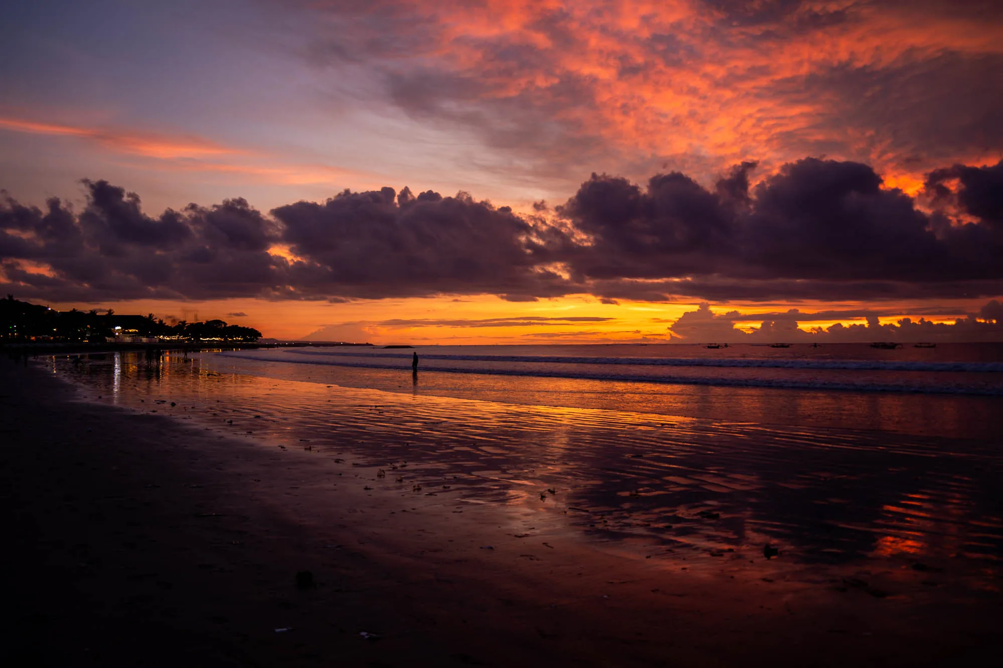 インドネシア、バリ島のビーチで撮影された夕焼けの風景写真です。手前の砂浜は、夕焼けの色を反射して赤黒く輝いています。中央には波打ち際が広がり、その先に水平線が見えます。水平線付近の海面には、いくつかの小舟が浮かんでいます。空には、オレンジ色や赤色に染まった雲が広がっており、ドラマチックな夕焼けを演出しています。雲の隙間からは、夕日の光が差し込み、幻想的な雰囲気を醸し出しています。遠くの海岸線には、明かりが灯った建物が並んでおり、静かな夜の始まりを感じさせます。全体的に、美しく、穏やかで、少し物憂げな印象を与える写真です。