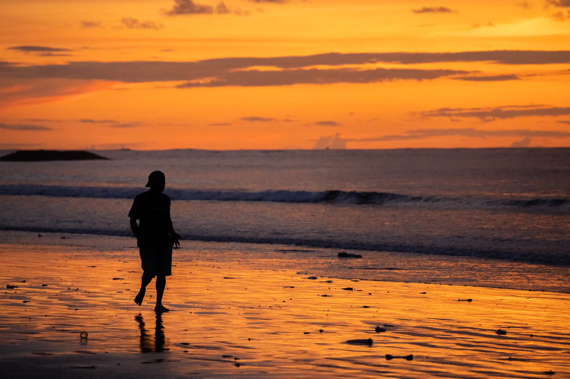 インドネシアのバリで撮影された写真です。画面全体は夕焼けのオレンジ色に染まり、水面がそれを反射して輝いています。手前には海岸線が広がり、濡れた砂浜は鏡のように空の色を映し出しています。砂浜には小さな漂流物や石が散らばっています。  中央やや左には、帽子をかぶった人物が半袖シャツと短パン姿で裸足で歩いているシルエットが見えます。人物の影も水面に長く伸びています。背景には穏やかな波が打ち寄せる海が広がり、水平線上に小さな島影が見えます。空には雲が点在し、夕焼けのグラデーションを際立たせています。全体的に静かで穏やかな、美しい夕暮れの風景を捉えた写真です。