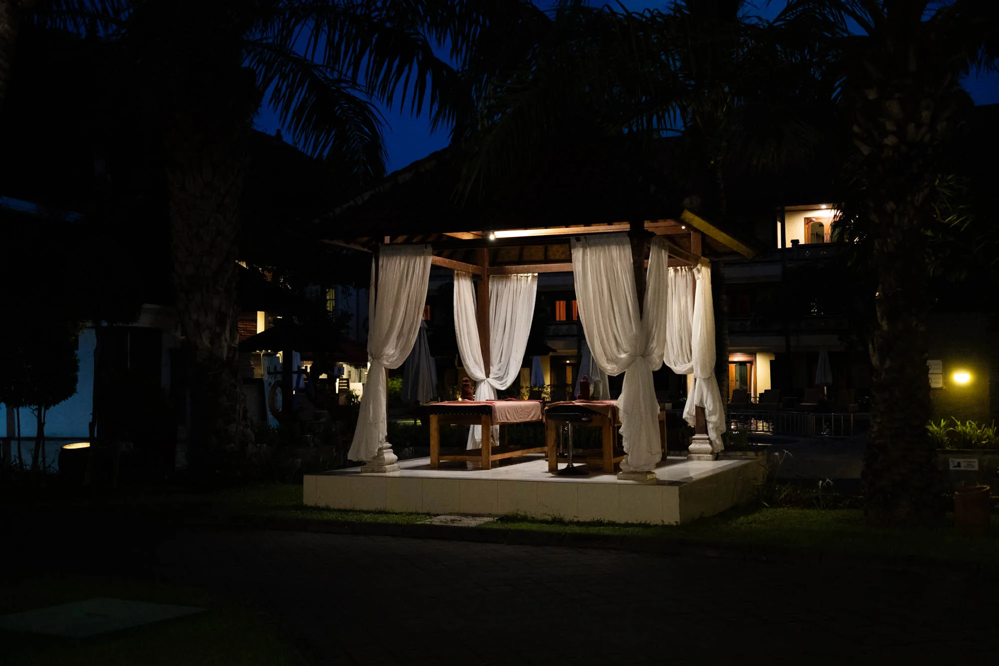 写真はインドネシアのバリ島で撮影されたと思われる、夜の風景です。薄暗い中、ライトアップされたガゼボが中心に配置されています。ガゼボは木製のフレームで、白いカーテンが風に揺れるように飾られています。内部にはマッサージベッドが2台並んでおり、静かでリラックスできる空間を演出しています。  周囲はヤシの木や熱帯植物に囲まれており、バリ島の夜らしい落ち着いた雰囲気を醸し出しています。背景にはホテルのような建物が見え、窓から漏れる光が温かさを添えています。全体的に暗いトーンで、ライトの光が強調され、幻想的な雰囲気を醸し出しています。静寂の中で、マッサージを受ける人にとって、特別な時間を提供してくれるような印象を受けます。