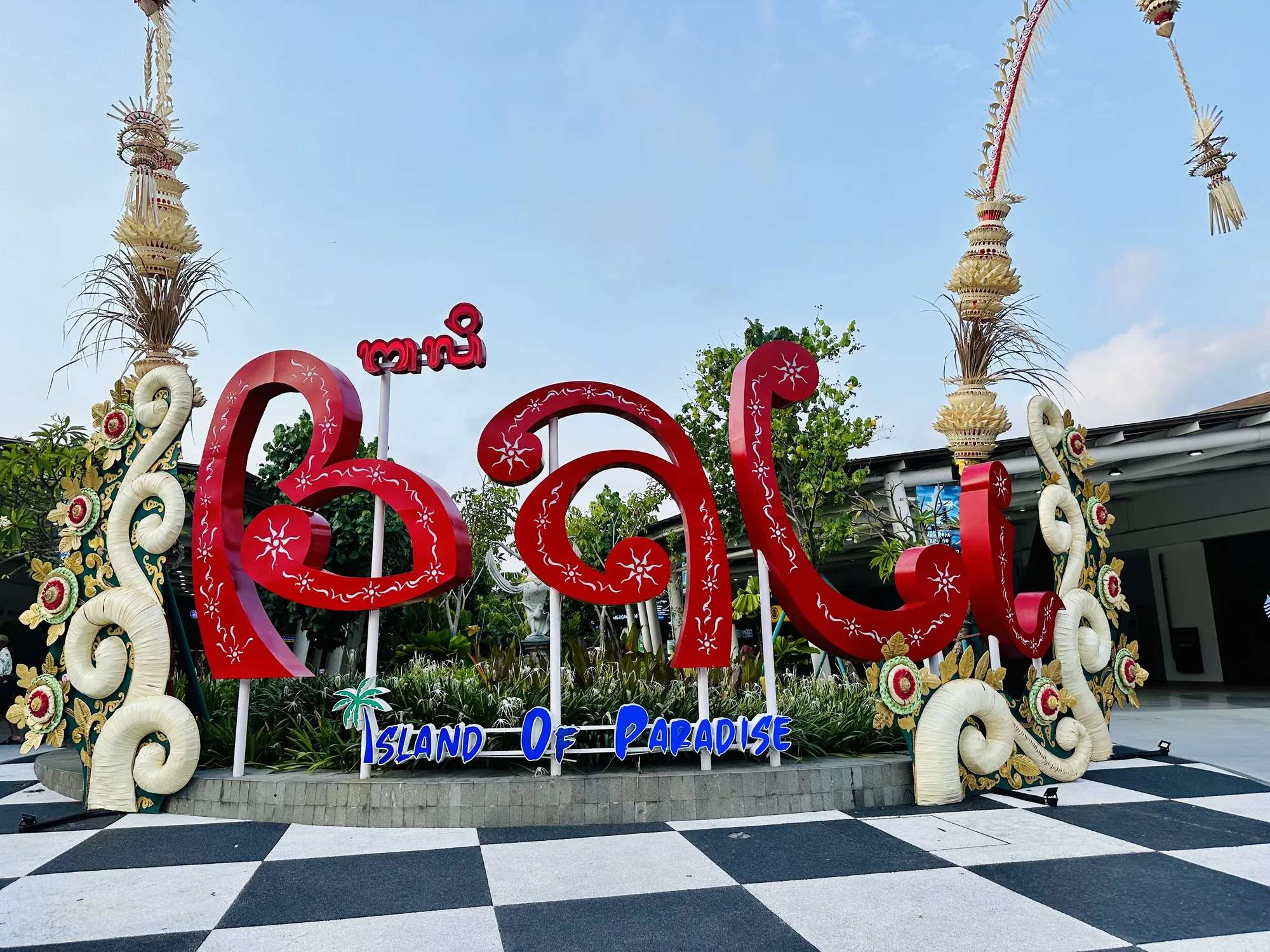 この写真はインドネシアのバリ島で撮影されたもので、観光客を迎えるためのモニュメントが写っています。モニュメントは、赤色で装飾された「Bali」の文字と、その下に青色で「ISLAND OF PARADISE」と書かれた看板で構成されています。文字はバリの伝統的な装飾が施されており、太陽のような模様が描かれています。モニュメントの両側には、バリの伝統的な装飾品である「Penjor（ペンジョール）」のようなものが飾られています。地面は黒と白のチェッカー柄で、現代的な雰囲気と伝統的な雰囲気が組み合わさったデザインです。背景には、緑豊かな植物と建物が見え、青空が広がっています。全体的に、バリ島の美しい自然と文化を象徴するような、歓迎ムードあふれる写真です。