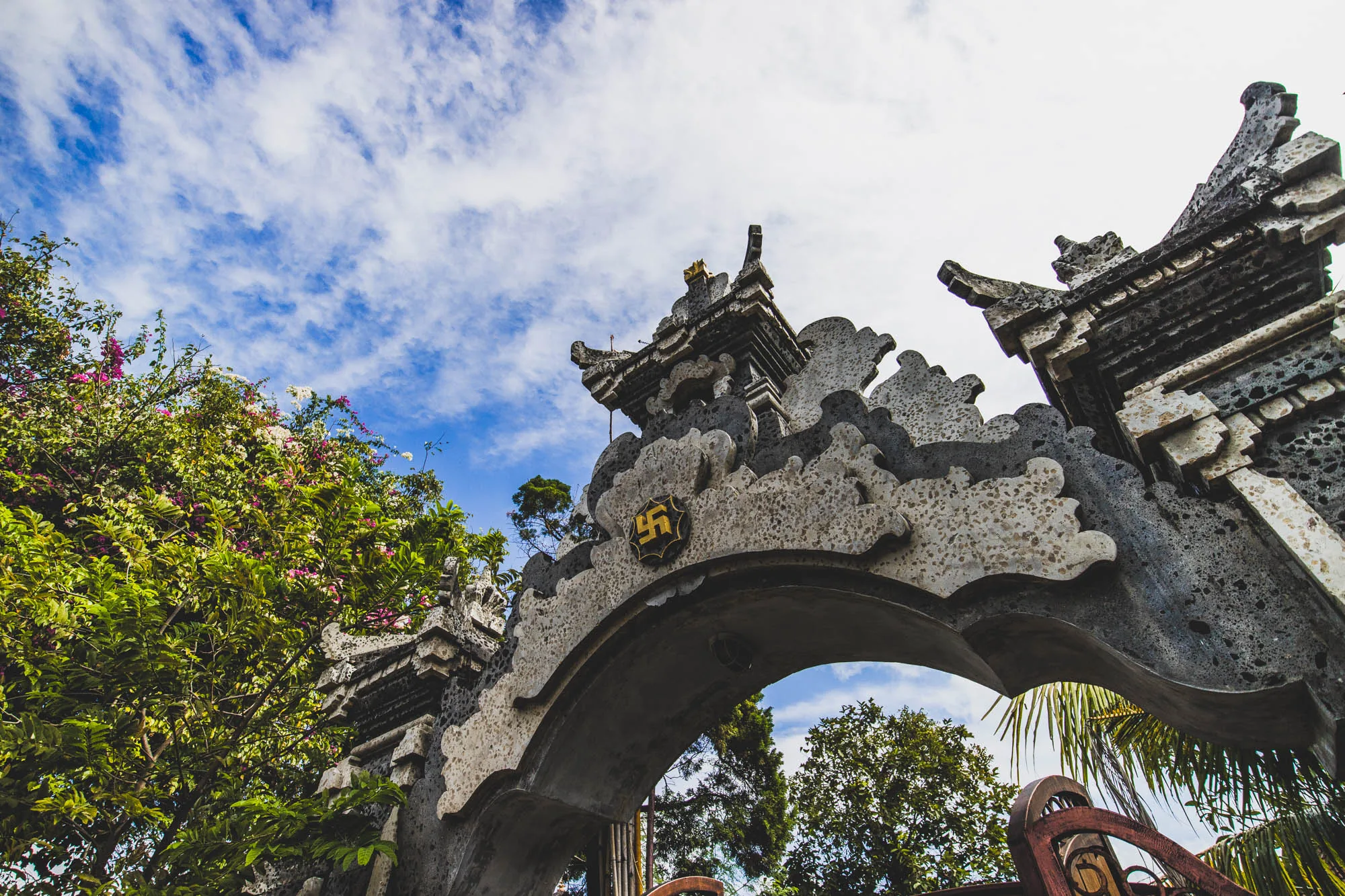 インドネシアのバリで撮影された写真です。写真は、伝統的なバリ様式の石造りの門をローアングルから捉えています。門は複雑な彫刻が施されており、ところどころに苔が生えており、歴史を感じさせます。門の上部には、金色の装飾が施されたエンブレムが見られます。門の向こうには、青空と白い雲が広がり、その下には緑豊かな木々が見えます。門の左側には、ブーゲンビリアなどの花が咲き乱れる木々が茂っています。写真は、バリの伝統文化と自然の美しさを捉えた一枚です。