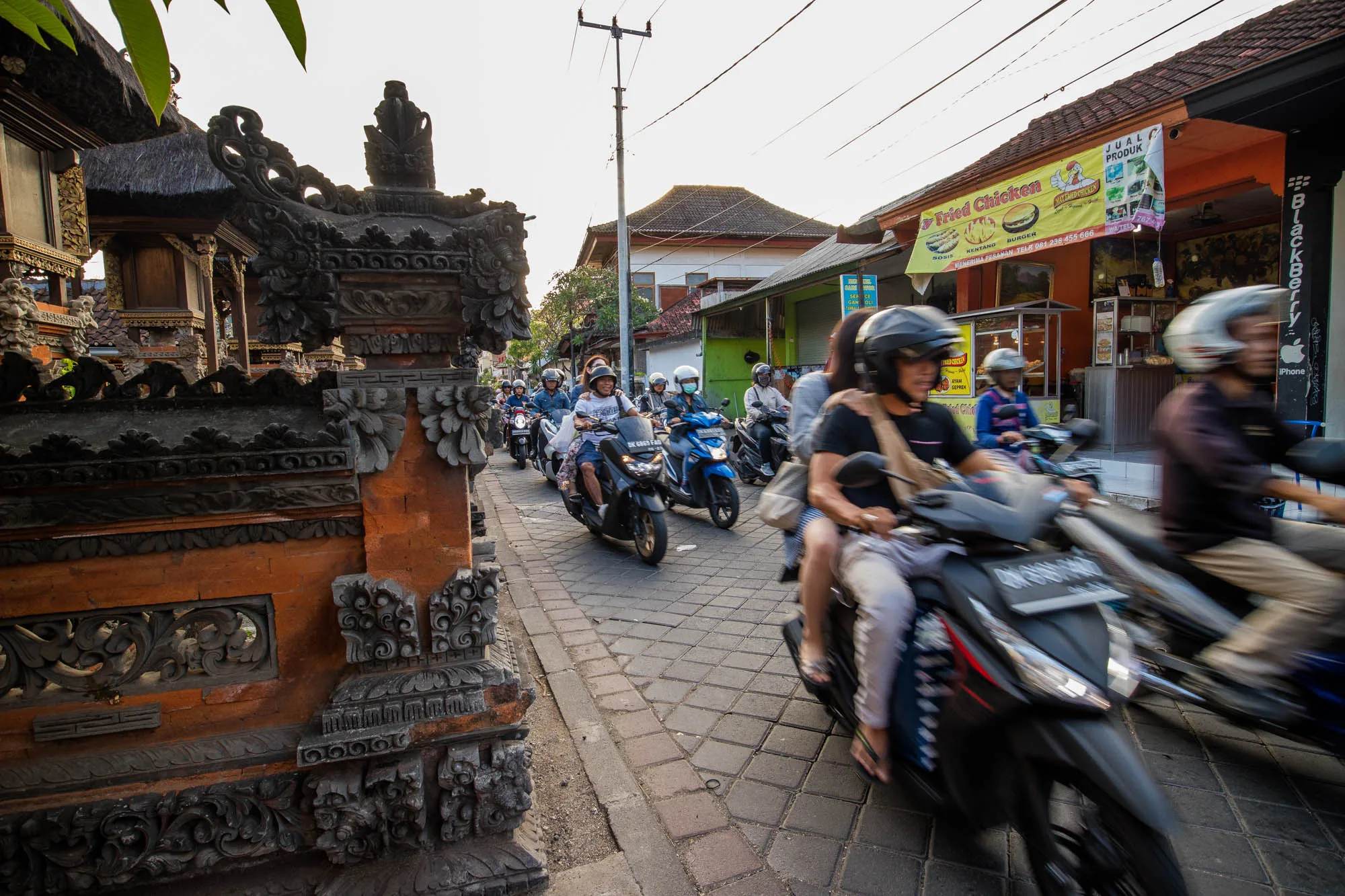 インドネシアのバリ島で撮影された写真です。石畳の狭い道路を、多くの人がバイクに乗って走っています。道路脇には、バリ独特の装飾が施された伝統的な建物や、小さな商店が見られます。特に、写真右側には「Fried Chicken」と書かれた黄色い看板のお店があり、ローカルな雰囲気を醸し出しています。バイクに乗っている人々の服装は様々で、ヘルメットを着用している人もいれば、そうでない人もいます。全体的に、活気があり、日常の風景を切り取ったような印象の写真です。