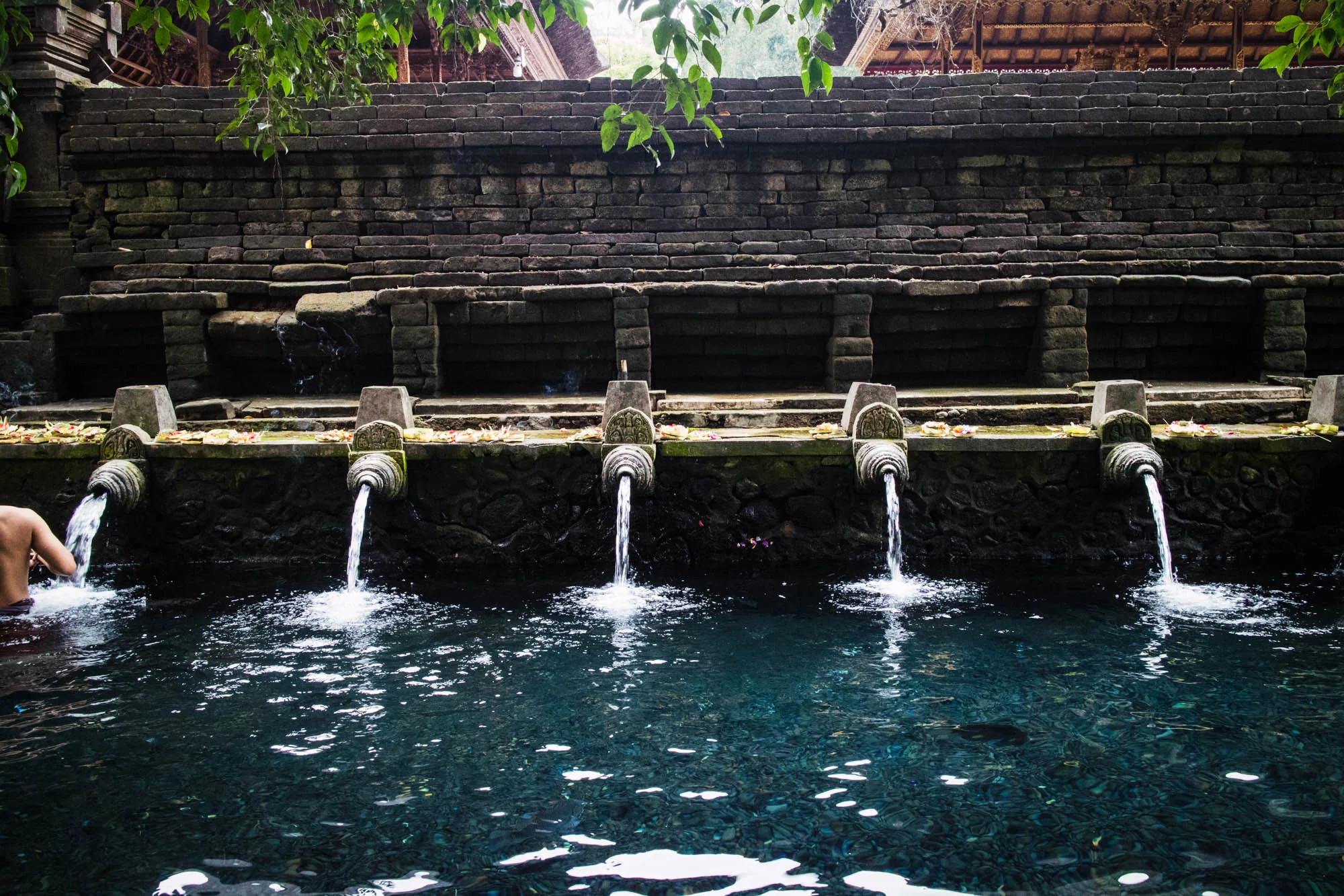写真はインドネシア、バリ島で撮影された聖なる泉の様子を捉えています。画面奥には、苔むした石造りの壁があり、石段のような構造になっています。壁面には複数の水口が設けられ、それぞれの水口から清水が流れ落ち、青く澄んだ水面に波紋を描いています。水口の上には、花びらのようなものが散りばめられています。左端には、水に浸かっている人の腕が見えます。背景には、伝統的な屋根を持つ建物の一部が確認できます。全体的に落ち着いた雰囲気で、神聖な場所の静けさと清涼感が伝わってきます。