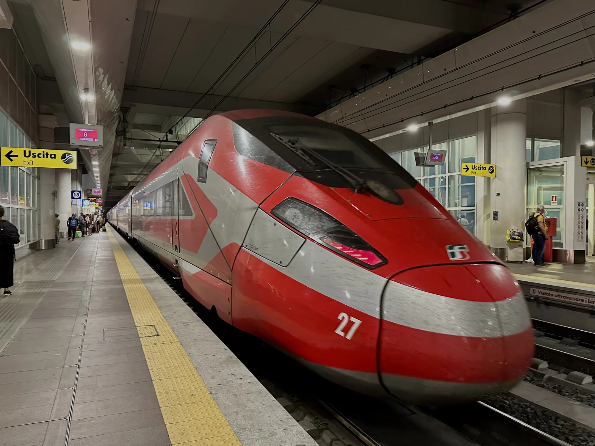 写真は、イタリアのフィレンツェにある駅のホームで撮影されたものです。画面中央には、赤とグレーのツートンカラーの高速列車が停車しています。列車の先頭部分が大きく写っており、「27」という数字が描かれています。列車の向こう側には、プラットフォームで待つ人々が見えます。頭上には出口を示す「Uscita Exit」の標識や、出発時刻を表示するモニターが設置されています。プラットフォームの床はグレーのタイルで舗装され、黄色い点字ブロックが敷かれています。全体的に、駅の喧騒と高速列車のモダンな雰囲気が伝わってくる写真です。