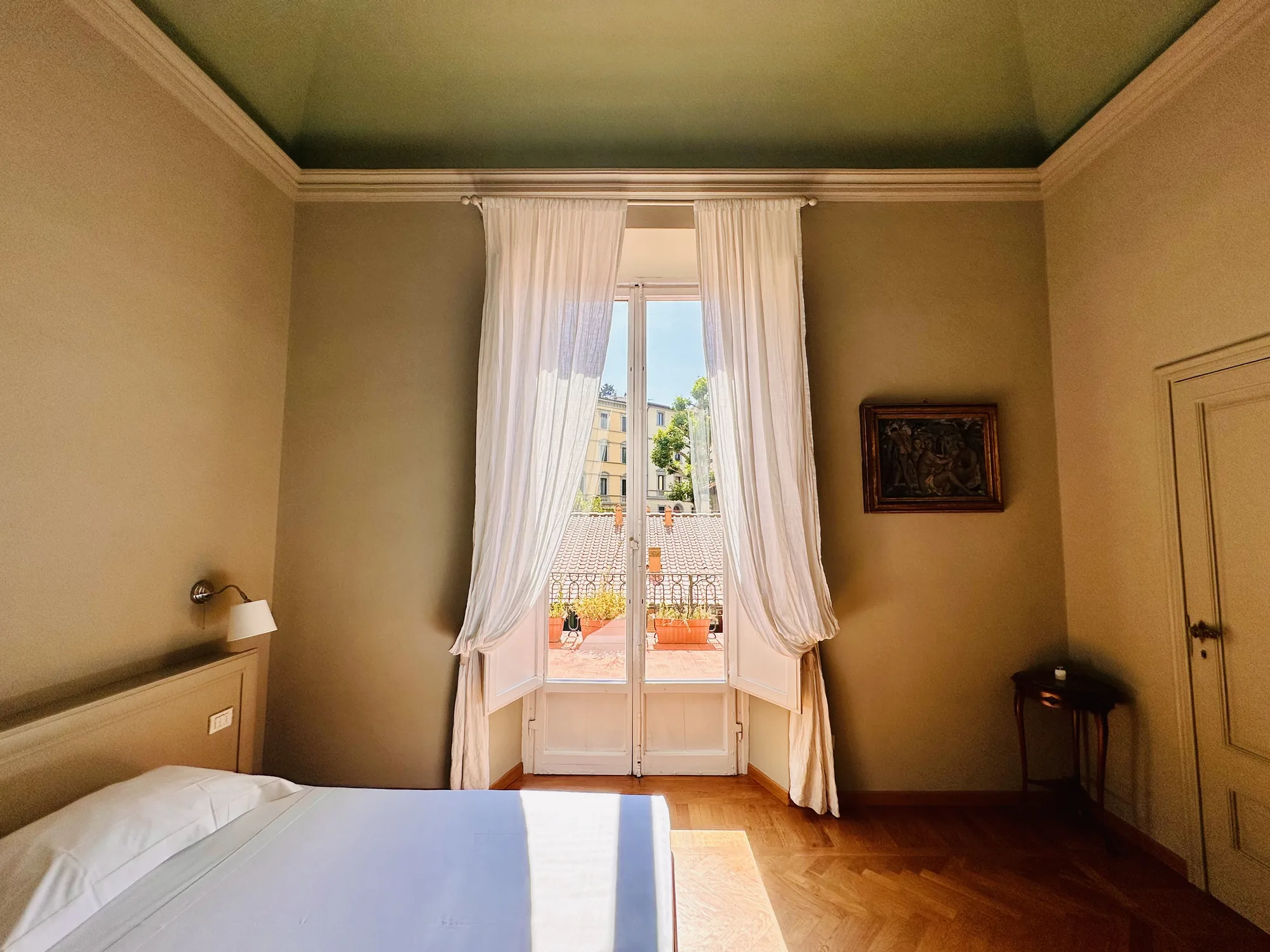 写真は、イタリアのフィレンツェで撮影された寝室の内部の様子を捉えています。部屋全体は暖色系の色調で統一されており、落ち着いた雰囲気を醸し出しています。画面左側にはベッドの一部が写っており、白いシーツが清潔感を漂わせています。ベッドの奥には壁に取り付けられた照明器具が見えます。  部屋の中央には、開放された白い窓があり、薄手のカーテンが柔らかく光を遮っています。窓の外には、テラコッタ色の屋根やバルコニー、そして緑豊かな木々が写っており、フィレンツェの美しい街並みが垣間見えます。窓から差し込む光が、部屋の床に暖かく射し込んでいます。  画面右側の壁には、額装された絵画が飾られています。絵画の内容は詳細には分かりませんが、人物が描かれているようです。絵画の下には、小さなテーブルとドアが配置されており、部屋の奥行きを感じさせます。  部屋の天井は淡い緑色で塗られており、壁との境には白いモールディングが施されています。床は木製の寄木細工で、光沢があり、上品な印象を与えています。全体的に、伝統的なイタリアの家屋の趣を感じさせる、快適で落ち着いた空間が表現されています。