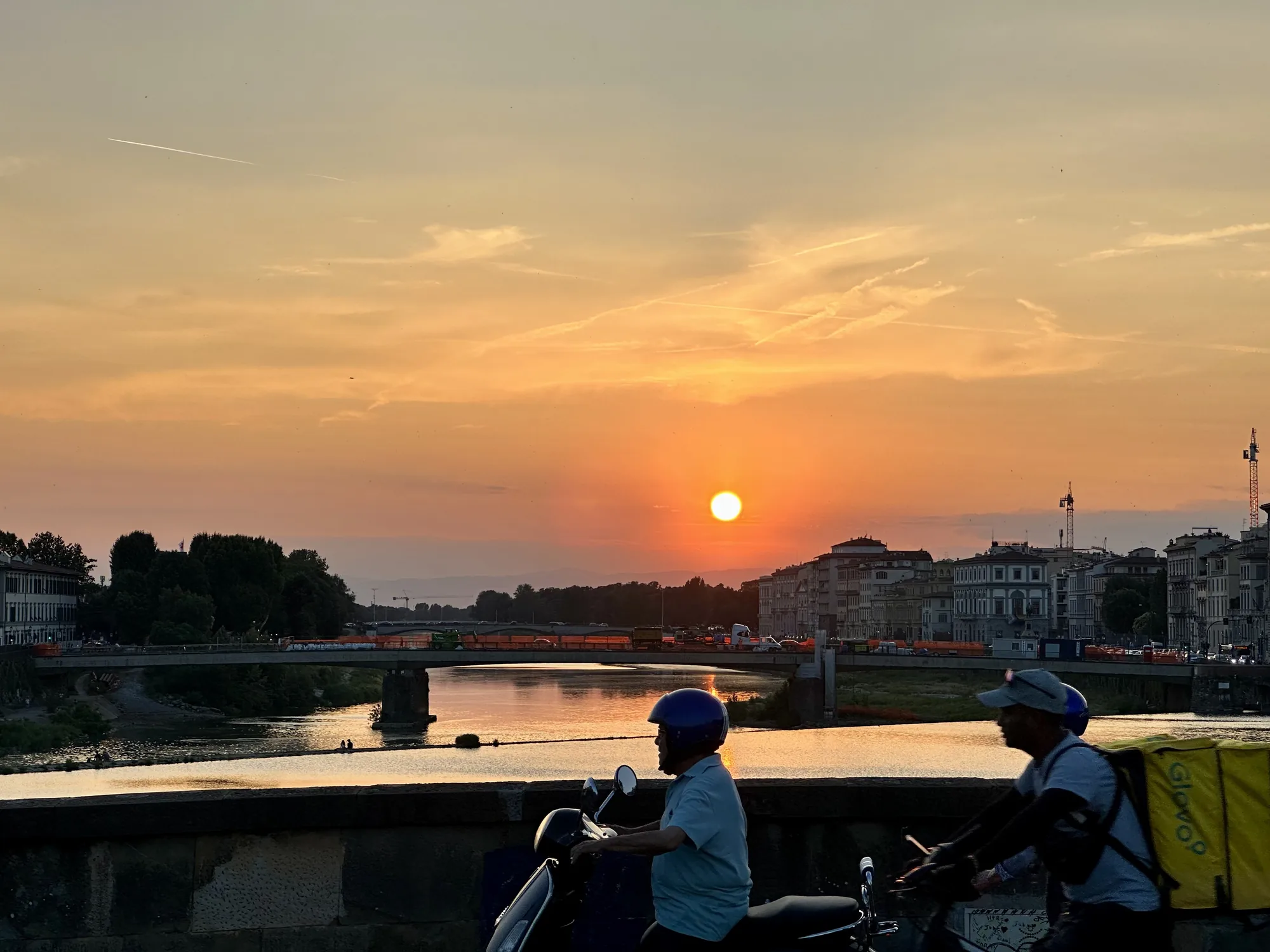 イタリアのフィレンツェで撮影された、夕暮れ時の風景写真です。太陽が地平線に沈みかけ、空はオレンジ色とピンク色のグラデーションに染まっています。空には飛行機雲が一本走っています。画面中央には、アルノ川がゆったりと流れ、川面は夕日に照らされて金色に輝いています。川には橋が架かっており、橋の上を車が走っているのが見えます。川沿いには歴史的な建物が立ち並び、フィレンツェらしい景観を形作っています。  前景には、橋の手すりの一部と、2台のバイクが写っています。手前側のバイクに乗る男性は、青いヘルメットを被っています。奥側のバイクに乗る男性は、Glovoの配達用リュックを背負っています。配達員が夕暮れの街を走る様子が、日常の風景を切り取ったかのような印象を与えます。全体的に暖かく穏やかな雰囲気の写真です。
