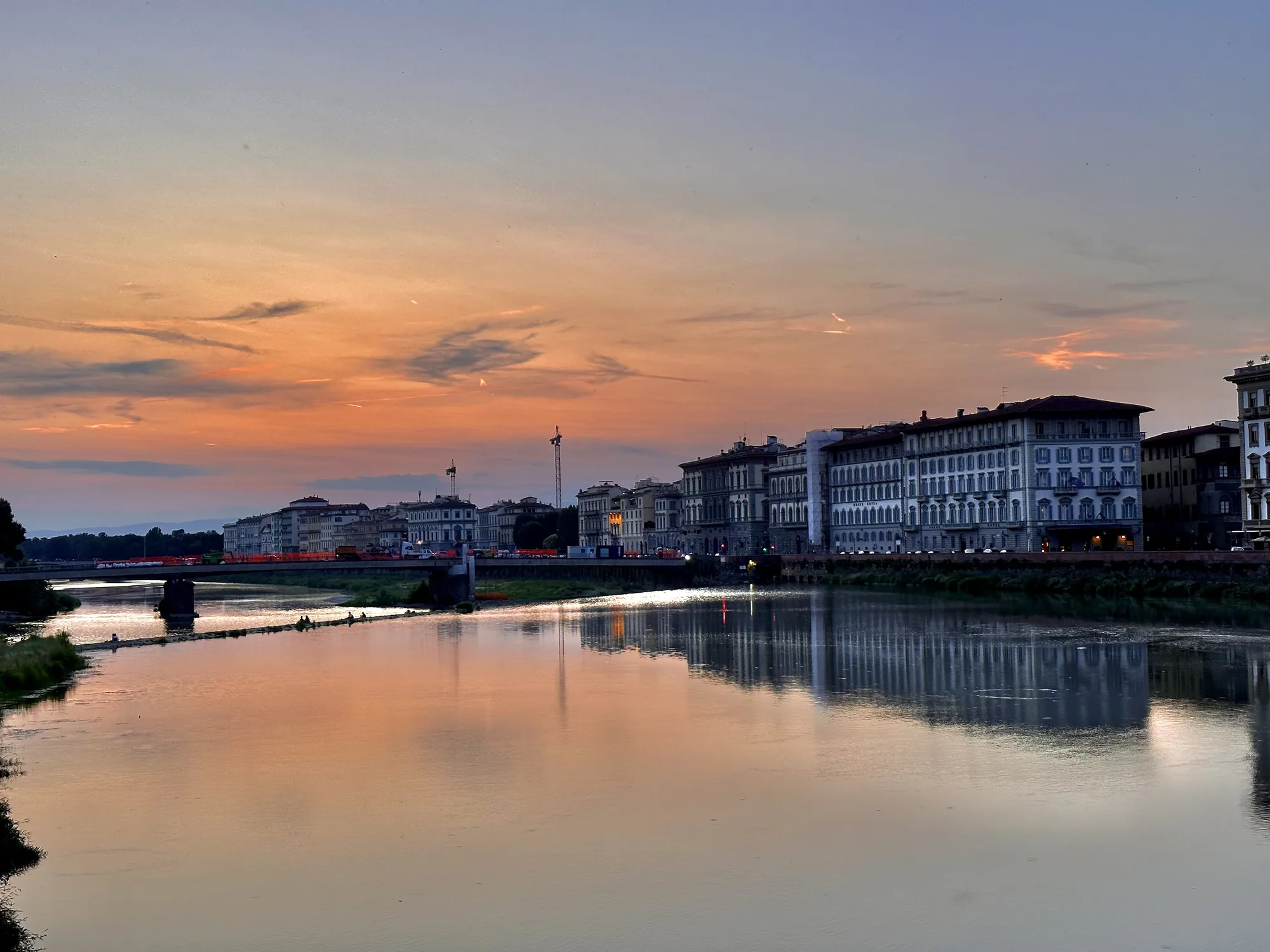 イタリアのフィレンツェで撮影された夕暮れ時の写真です。画面中央にはアルノ川が広がり、穏やかな水面には夕焼け空の色が反射して、オレンジ色に染まっています。川の奥には橋が架かっており、橋の向こうにはフィレンツェの街並みが続いています。建物の窓や街灯の明かりが水面に映り込み、幻想的な雰囲気を醸し出しています。空はグラデーションになっており、地平線に近い部分は濃いオレンジ色、上空に向かうにつれて薄い紫色に変化しています。夕暮れ時の静けさと美しさが感じられる一枚です。