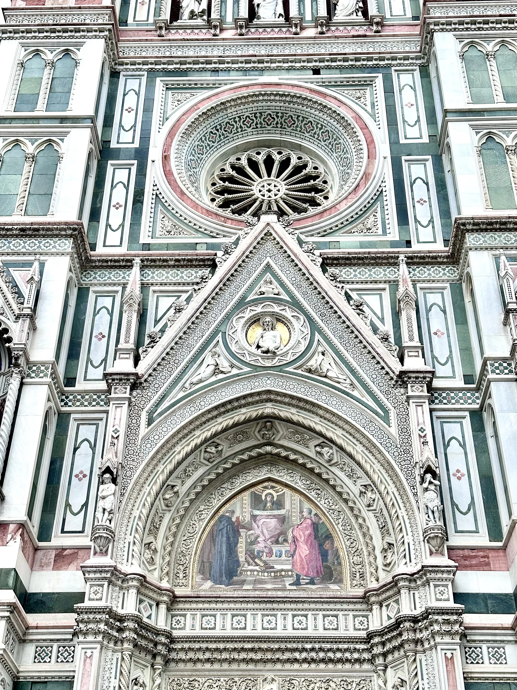イタリアのフィレンツェにあるフィレンツェ大聖堂（ドゥオーモ）の一部を捉えた写真です。写真は建物の正面入り口上部をクローズアップしており、その精緻な装飾が際立っています。白、緑、ピンクの大理石を組み合わせた幾何学的なパターンが特徴的で、ゴシック様式の窓やバラ窓、彫刻、モザイク画が細部にまで施されています。入り口上部には聖母マリアを描いたと思われる絵画が見られ、その周囲を天使や人物の彫刻が飾っています。全体的に、建物の壮大さと芸術的な美しさを感じさせる一枚です。