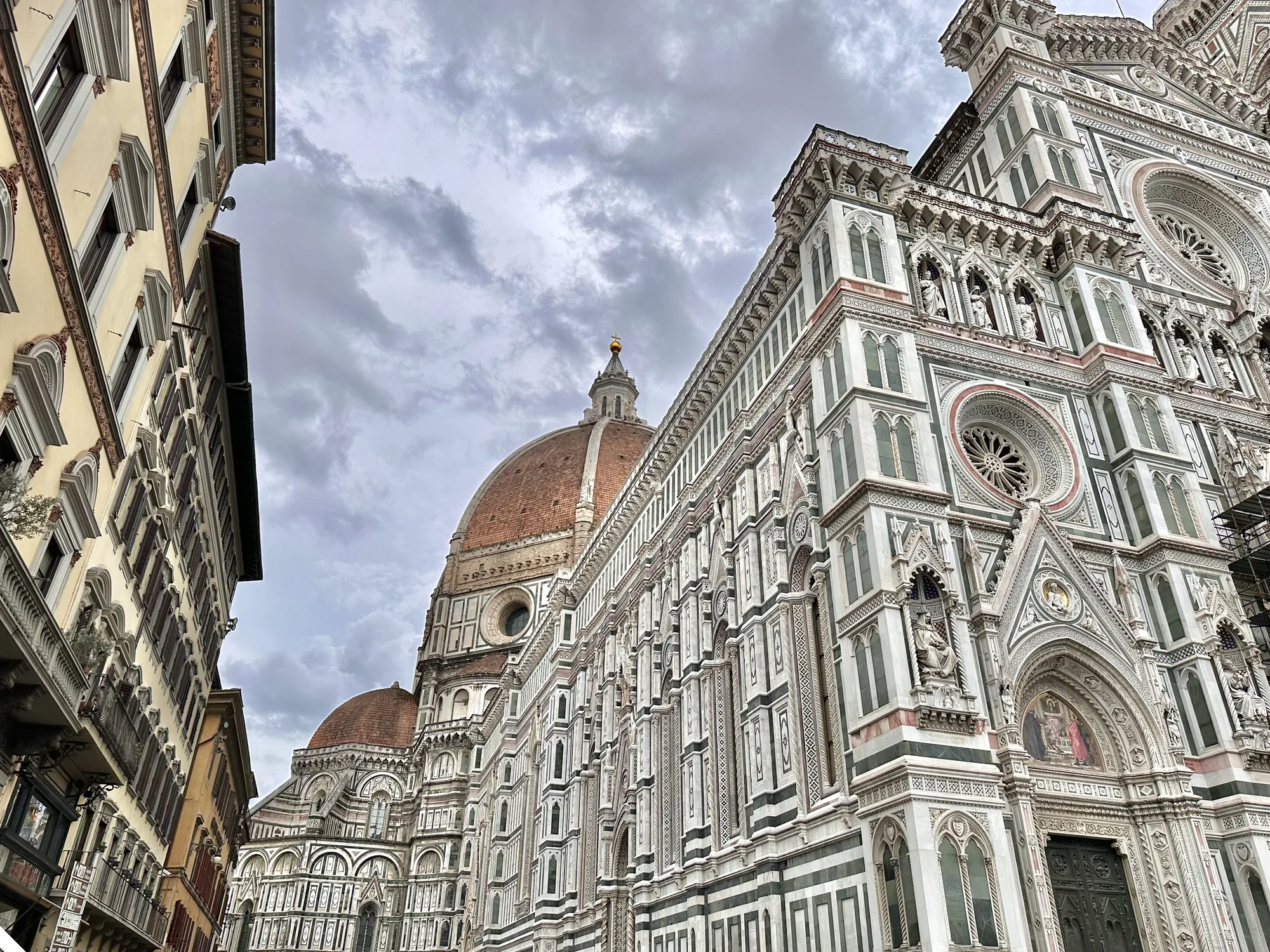 イタリアのフィレンツェで撮影された写真です。画面中央から右側にかけて、壮大なフィレンツェのドゥオーモ（サンタ・マリア・デル・フィオーレ大聖堂）がそびえ立っています。ドゥオーモは、白を基調とした外壁に緑色の装飾が施され、複雑な彫刻や装飾が目を引きます。建物の細部まで丁寧に作り込まれており、ルネサンス建築の美しさを際立たせています。  ドゥオーモの左側には、古い建物が並ぶ通りが見えます。建物の壁は黄色で、窓には茶色の鎧戸がついています。歴史を感じさせる建物が、フィレンツェの街並みを彩っています。  空は厚い雲に覆われており、ややどんよりとした天気です。しかし、雲間からわずかに光が差し込み、建物を幻想的に照らしています。全体的に落ち着いた雰囲気の写真ですが、ドゥオーモの存在感が圧倒的で、見る者を魅了します。