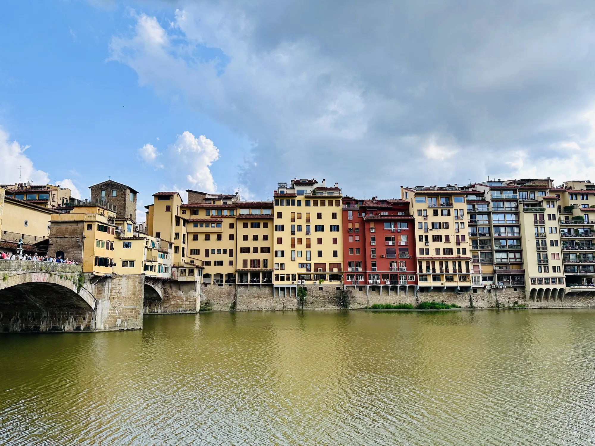 イタリアのフィレンツェで撮影された写真です。画面の中央には、穏やかな水面の川が広がっています。川の色はやや濁った黄緑色で、微かな波紋が全体に広がっています。川の向こう岸には、様々な色合いの建物が立ち並び、それぞれ異なる高さとデザインを持っています。建物の色は、薄い黄色、オレンジ色、赤色など様々で、窓の配置も不規則です。建物の一番手前には、石造りの橋が見え、橋の上には多くの人が歩いている様子が写っています。橋のアーチは、川面に影を落としています。空は青空と白い雲が混ざり合い、一部には灰色の雲も見られます。全体的に落ち着いた雰囲気の写真です。