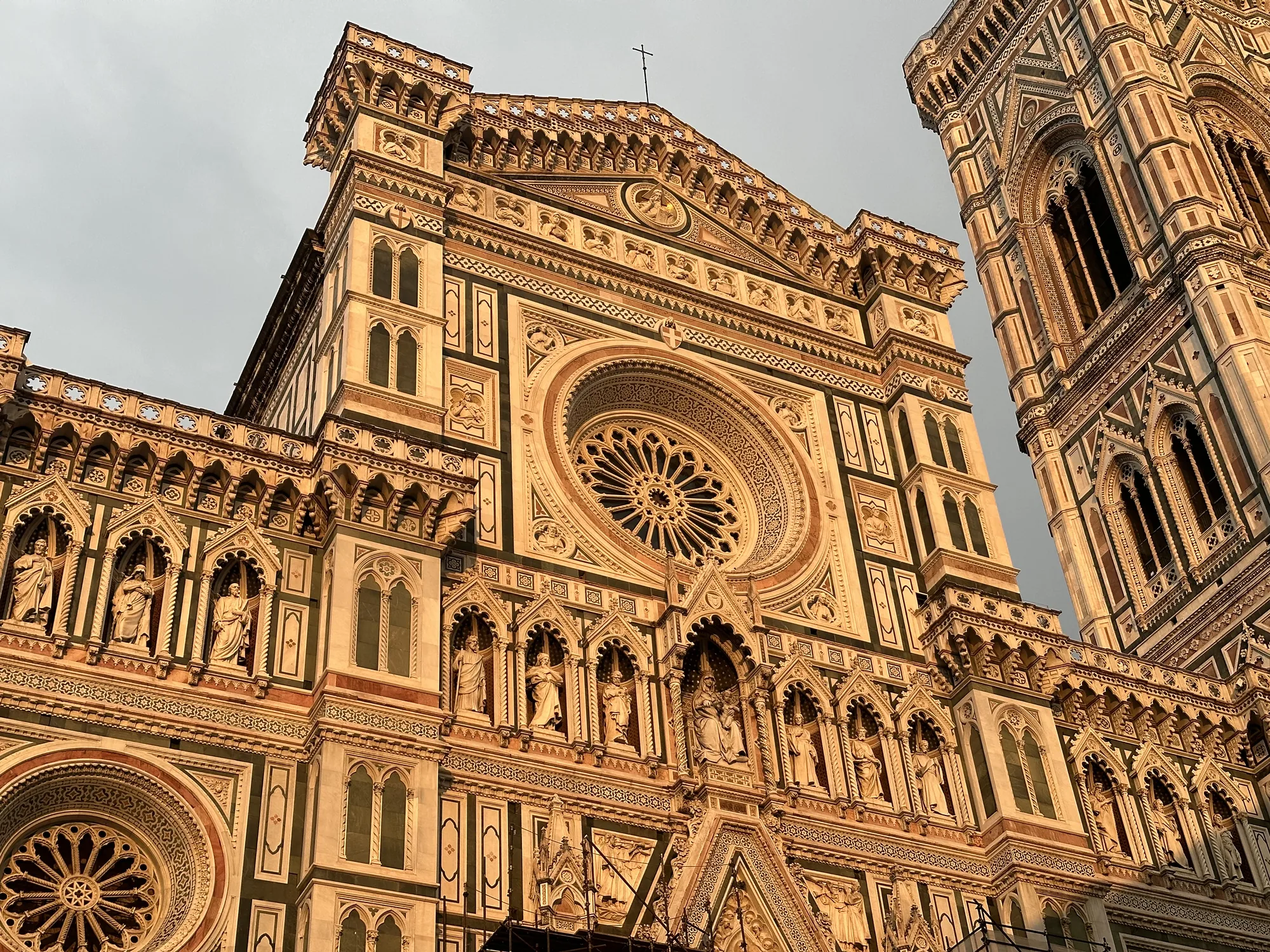 写真はイタリアのフィレンツェにあるフィレンツェ大聖堂（ドゥオーモ）を捉えたものです。ローアングルから見上げる構図で、建物の壮大さと精緻な装飾が際立っています。  建物の外壁は白、緑、ピンクの大理石で覆われており、複雑な幾何学模様と彫刻が施されています。写真には、精巧な彫刻が施された聖人像が多数並んでいるのが見えます。また、建物の正面には、大きなバラ窓（ロゼット窓）があり、その繊細な模様が目を引きます。建物の頂上には十字架が掲げられています。  写真全体は夕日のような暖かい光に照らされており、石造りの建物に陰影を作り出し、その重厚感を強調しています。空は曇っているように見えますが、光と影のコントラストが建物の美しさを際立たせています。写真の右側には、ジョットの鐘楼の一部が写っており、こちらも同様の美しい装飾が施されています。  全体的に、写真からはフィレンツェ大聖堂の歴史と芸術的な価値が伝わってきます。建物の細部まで丁寧に表現されており、見る者に感動を与えるような荘厳な雰囲気を醸し出しています。