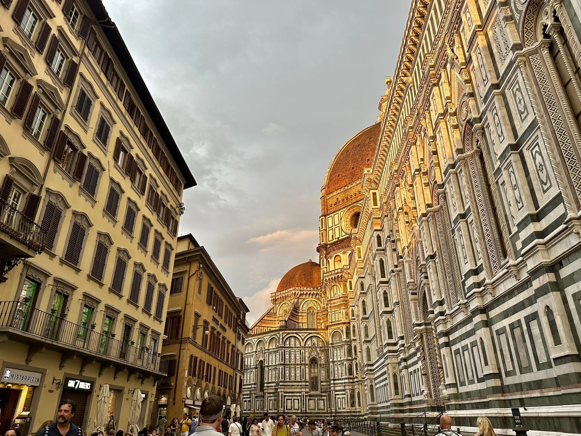 イタリアのフィレンツェで撮影された写真です。画面右側に壮大なフィレンツェ大聖堂（ドゥオーモ）の一部が大きく写っています。大聖堂の外壁は白と緑の模様で装飾されており、夕日の光を受けて暖かく輝いています。大聖堂のドームも一部見え、レンガ色のような赤茶色が印象的です。  画面左側には、窓がたくさんある黄色の建物がそびえ立っています。建物の窓には茶色の鎧戸が見えます。写真全体はローアングルで撮影されており、大聖堂や建物を見上げるような構図になっています。  写真の下部には、多くの人々が歩いている様子が写っています。観光客と思われる人々が、大聖堂を背景に写真を撮ったり、談笑したりしているようです。空は曇り空で、夕焼けのような色合いも見られます。全体的に、歴史的な建造物の美しさと、そこに集まる人々の賑わいが感じられる一枚です。