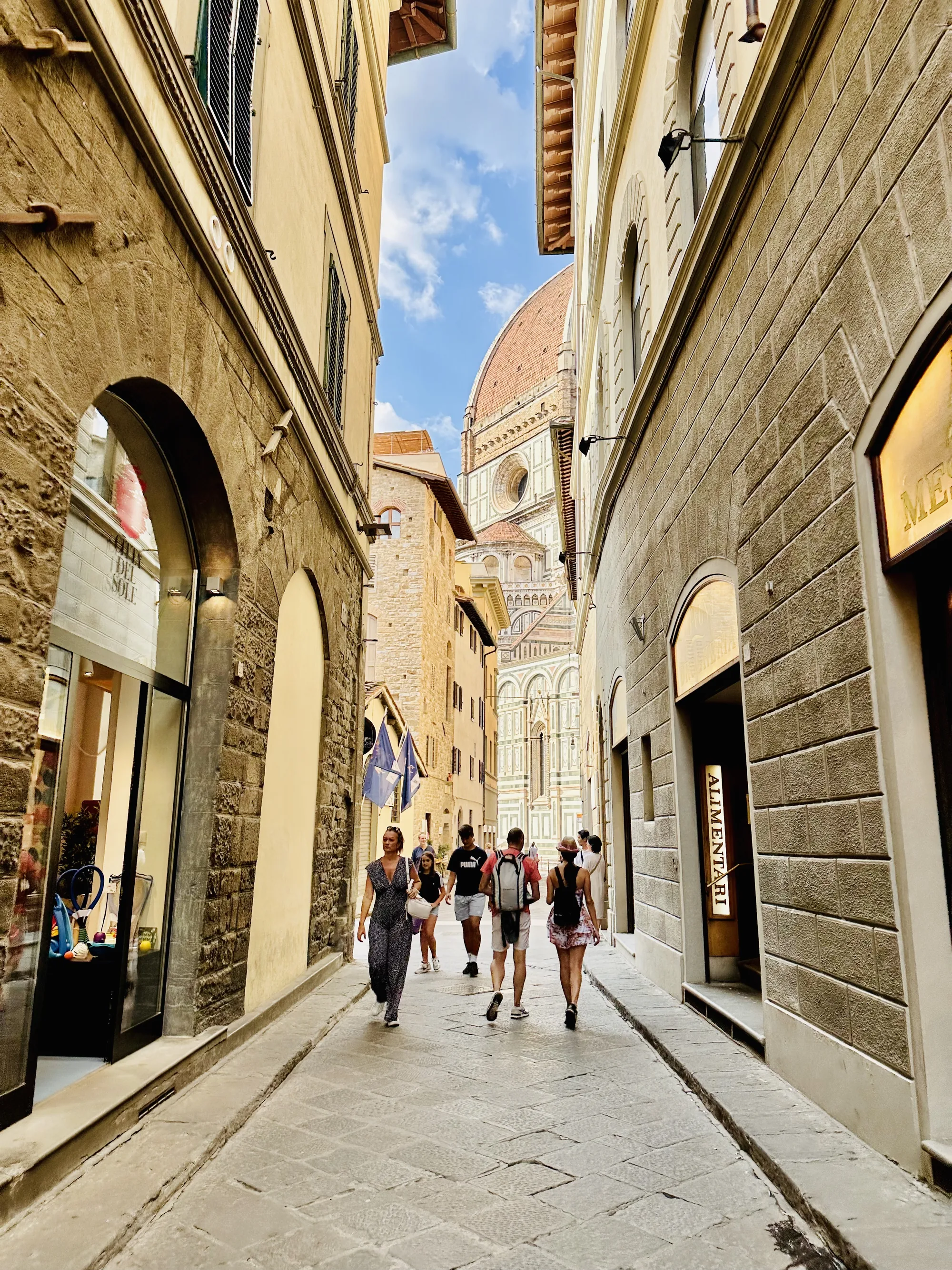 イタリアのフィレンツェで撮影された写真です。狭い路地の奥に、フィレンツェのドゥオーモの巨大なドームが見えています。路地の両側には石造りの建物が立ち並び、アーチ状の入り口や窓が歴史を感じさせます。路面の石畳が古い街並みの雰囲気を強調しています。何人かの観光客が路地を歩いており、観光地らしい賑わいを感じさせます。青い空と白い雲が背景に広がり、建物の色合いと相まって美しいコントラストを生み出しています。全体的に、落ち着いた色調で、歴史的な街並みを歩く人々の日常が写し出されています。