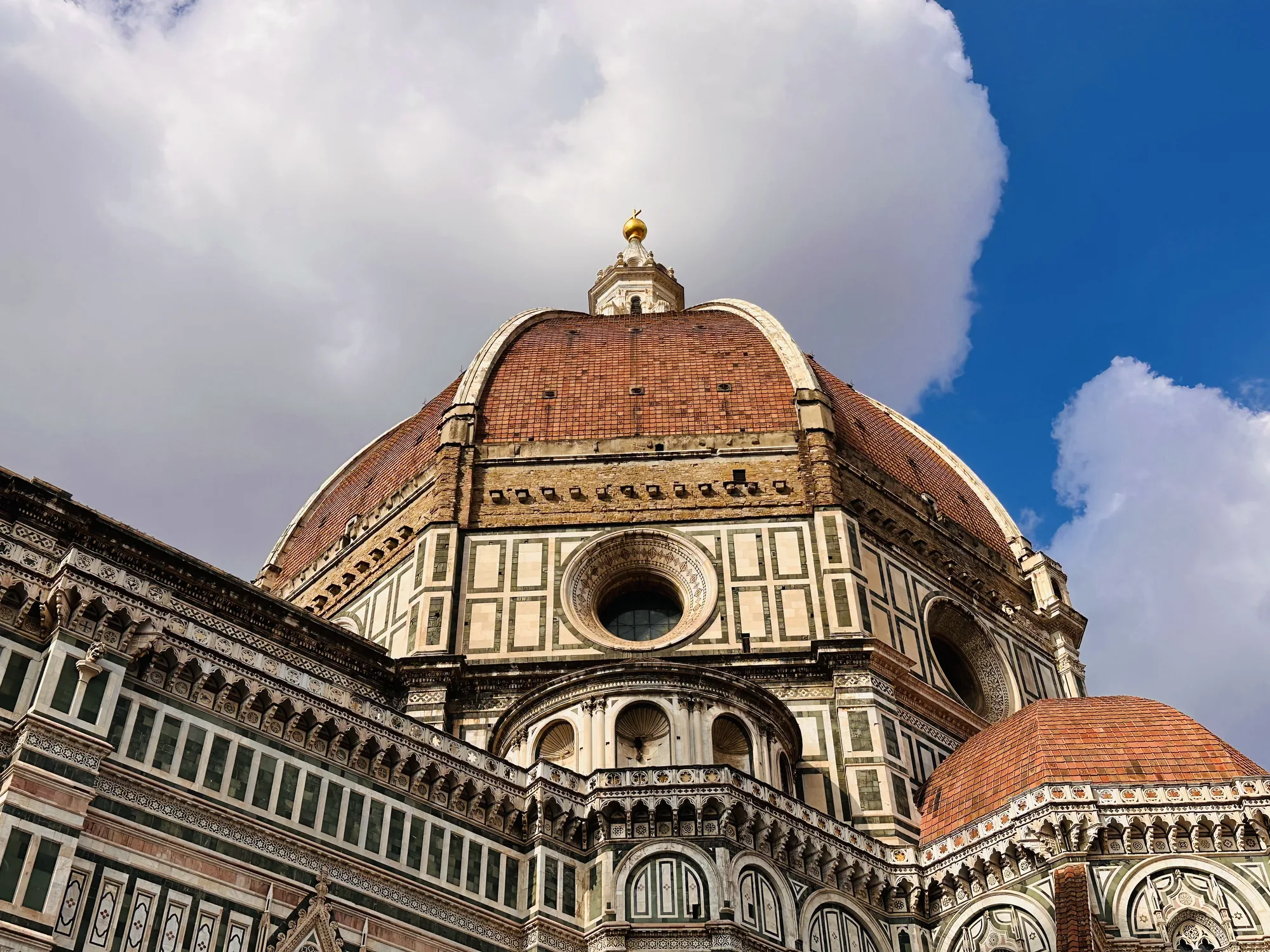 イタリアのフィレンツェにあるドゥオーモ、サンタ・マリア・デル・フィオーレ大聖堂のクーポラ（ドーム）を捉えた写真です。画面全体にドームが迫力ある構図で写っています。ドームの表面は赤茶色のレンガで覆われており、その下部には白と緑の大理石で装飾された美しい装飾が見られます。装飾には、幾何学的な模様や円形の窓、アーチなどが緻密に配置されており、ルネサンス建築の壮大さを感じさせます。ドームの頂上には、金色の球体と十字架が輝いており、青空に映えています。背景には白い雲が浮かび、明るい光がドーム全体を照らしています。