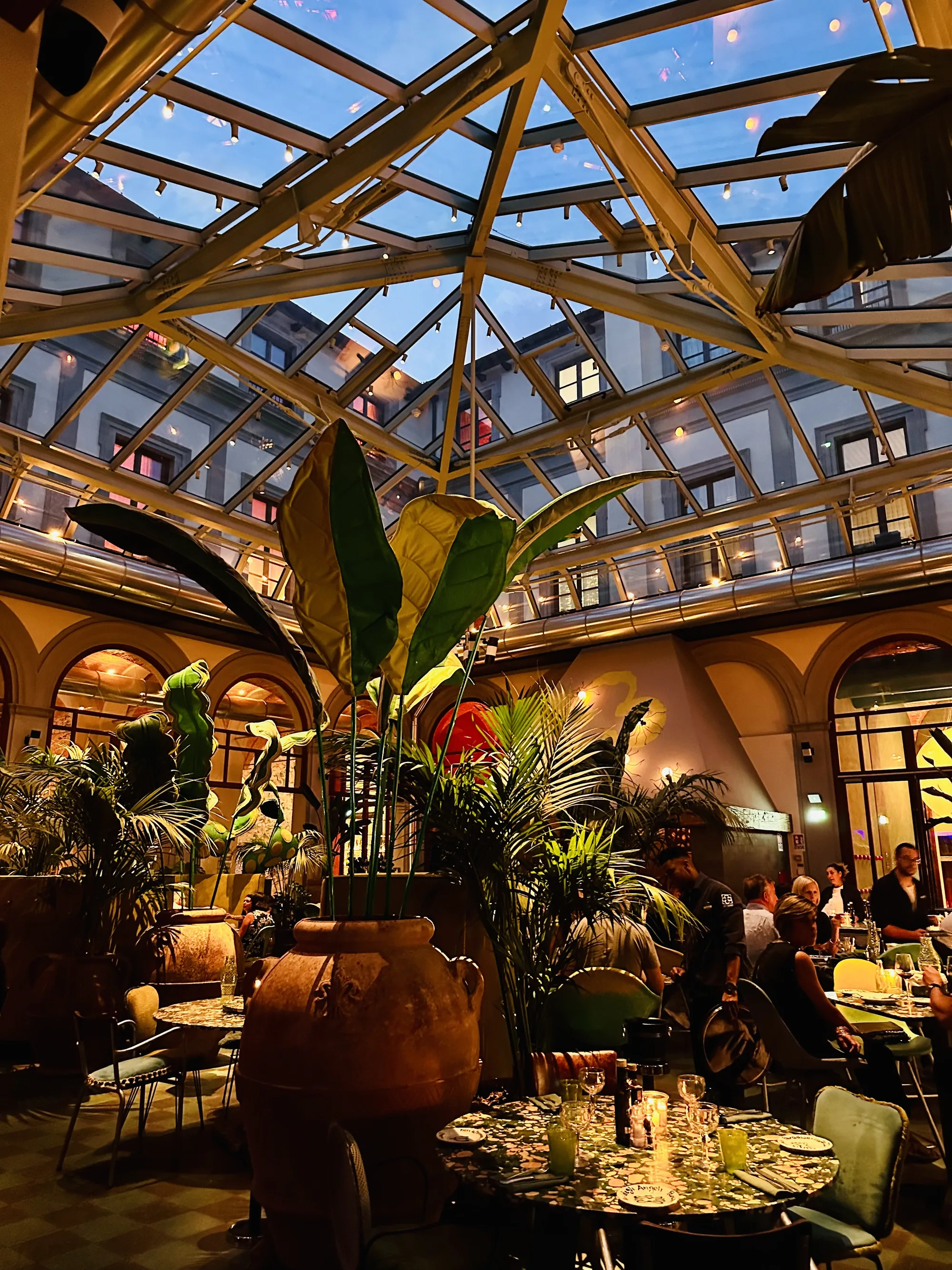 イタリアのフィレンツェで撮影された、ガラス屋根の温室のようなレストランの写真です。ガラス屋根からは夕暮れ時の空が見え、建物内部の照明が点灯しています。レストラン内はテーブル席が並び、人々が食事を楽しんでいます。テーブルの上にはキャンドルが灯され、温かみのある雰囲気を醸し出しています。大きな植木鉢に植えられた植物が空間を彩り、トロピカルな雰囲気を加えています。内装はアーチ状の窓や壁の装飾など、クラシックな要素も感じられます。全体的に落ち着いた照明で、リラックスした雰囲気のレストランであることがわかります。