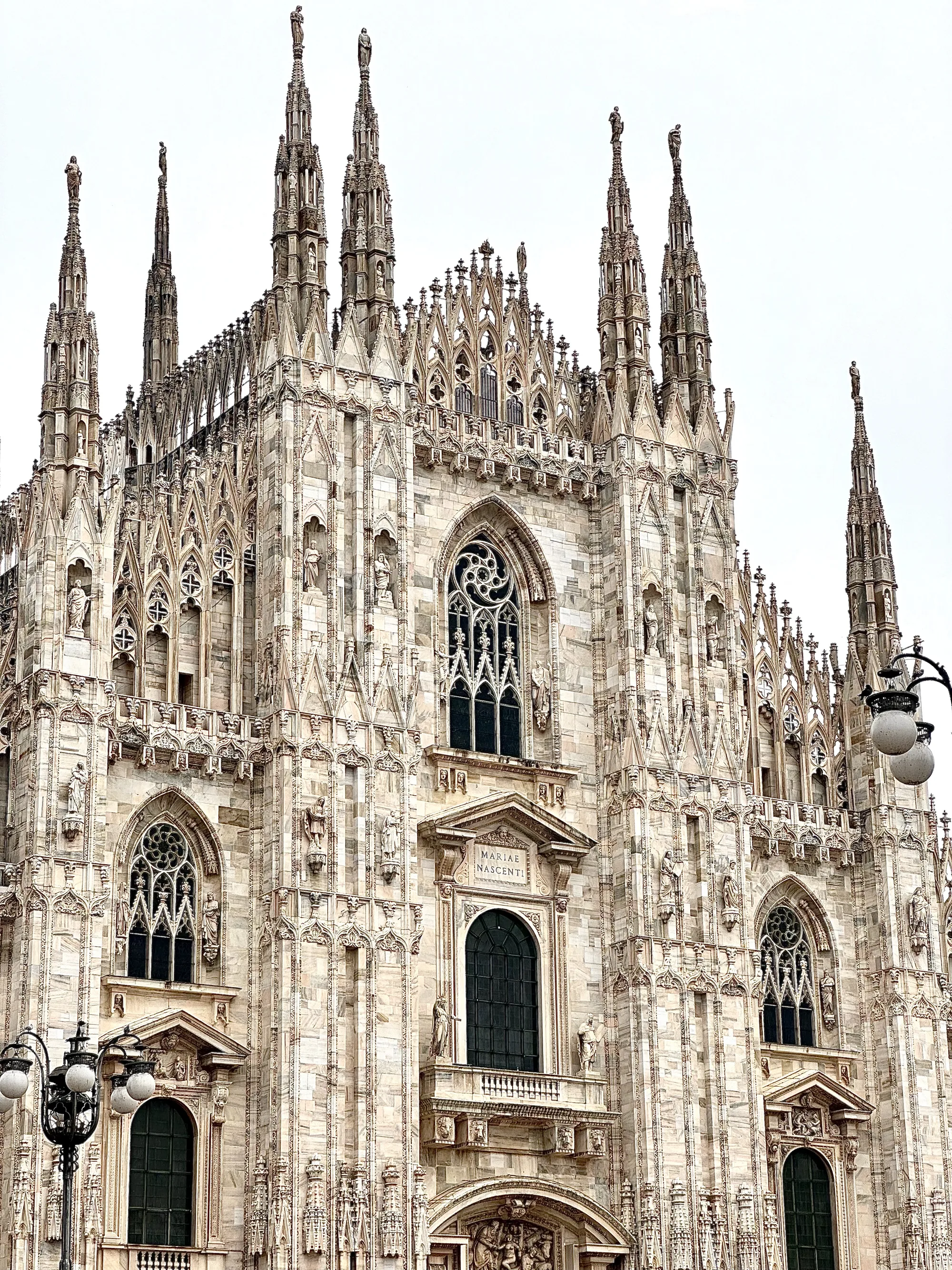 写真はイタリアのミラノにあるミラノ大聖堂の一部を捉えています。カメラは建物の正面を捉え、高い位置から見上げています。大聖堂の外観は非常に細かく、ゴシック様式の装飾が豊富です。尖塔がいくつも空に向かって伸び、それぞれに彫像が飾られています。壁面には無数の彫刻が施され、宗教的なテーマや人物を表しているようです。窓はアーチ型で、精巧なステンドグラスがはめ込まれています。写真全体は白っぽい色調で、空は曇っているように見えます。写真の左下には、街灯の一部が写り込んでおり、大聖堂のスケールをさらに強調しています。全体的に、この写真は大聖堂の壮大さと複雑な建築様式を印象的に伝えています。