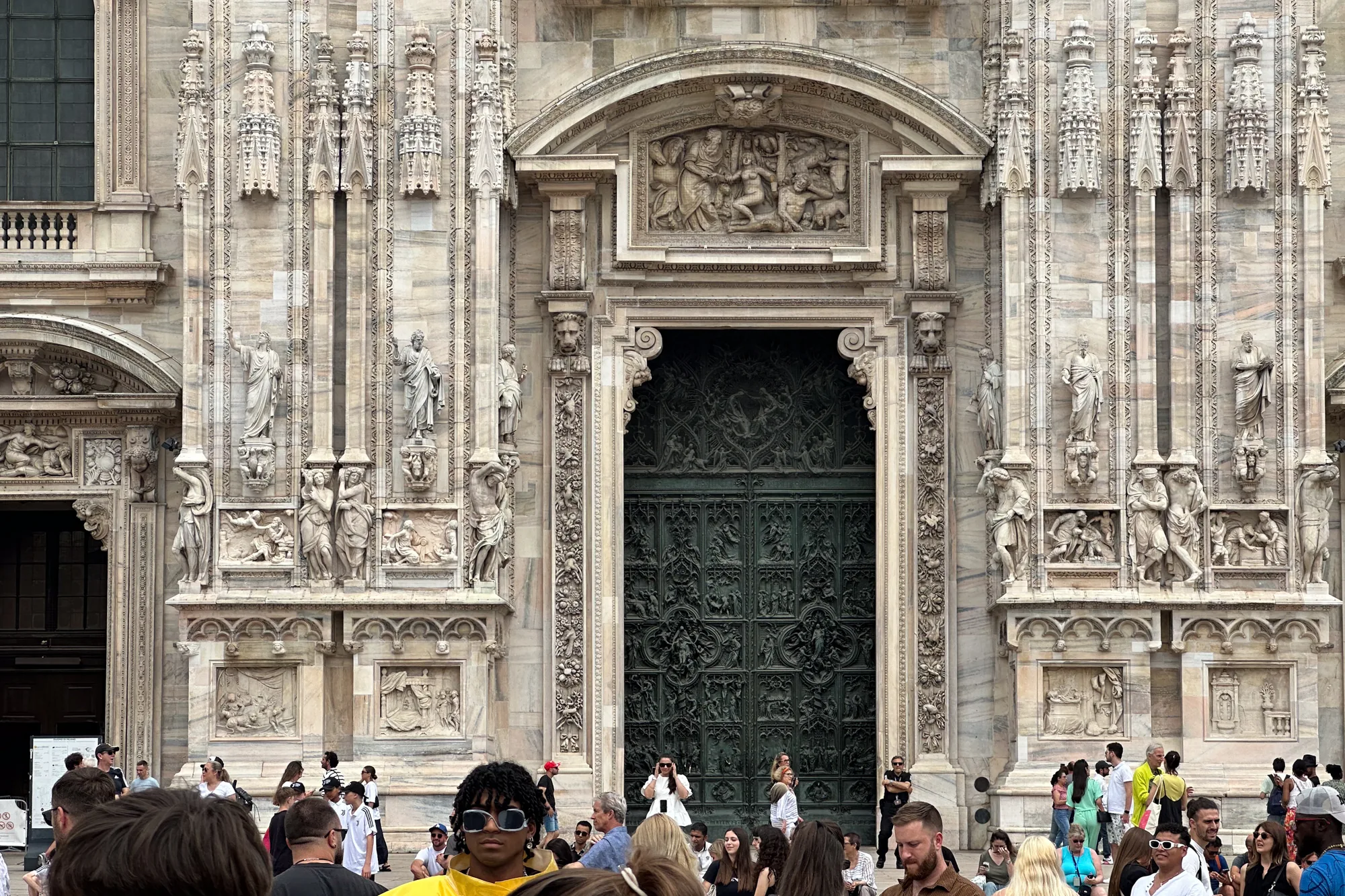 写真はイタリアのミラノで撮影されたもので、ミラノ大聖堂（ドゥオーモ）の壮大な扉を捉えています。扉は非常に細かく複雑な彫刻で覆われており、重厚感と歴史を感じさせます。扉の周囲は、大理石の壁面で飾られ、彫像やレリーフが施されています。壁面の石材は、美しい模様を描いており、建築の芸術性を高めています。  写真の下部には、大勢の観光客が写っています。様々な服装をした人々が、大聖堂の前に集まり、写真を撮ったり、談笑したりしています。観光客の存在は、この場所が人気の観光スポットであることを示しています。全体の雰囲気は、歴史的な建造物の荘厳さと、観光客の賑わいが組み合わさった、活気のあるものです。