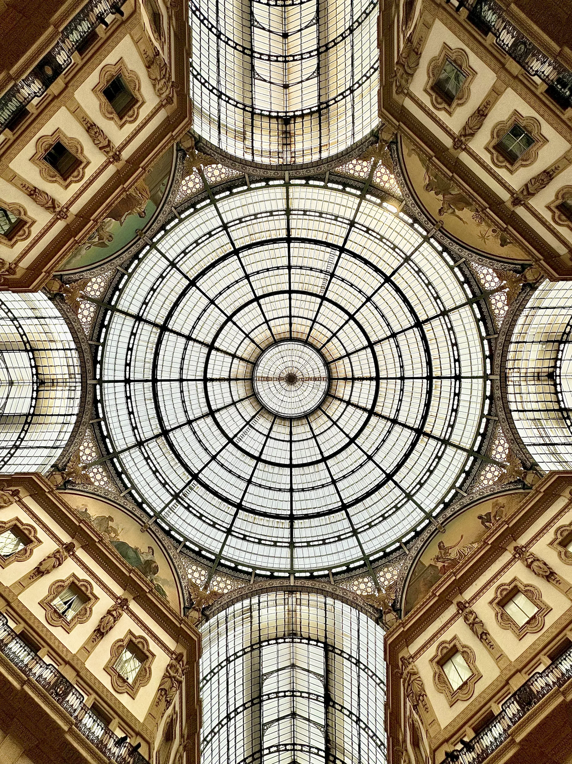 この写真はイタリアのミラノで撮影された、壮麗な天井の様子を捉えています。視点は真下から見上げるアングルで、中心には巨大なガラスドームが位置しています。ドームは幾何学的な模様を描くように金属のフレームで区切られており、光を透過して明るい空間を作り出しています。ドームを取り囲むように、建物の一部が見えています。壁面は淡いクリーム色で、窓や装飾が施されており、その細部までが丁寧に表現されています。壁面には美しい絵画が描かれ、建物の歴史と芸術性を物語っています。写真全体は、壮大さと繊細さが共存した、息をのむような美しさを感じさせます。