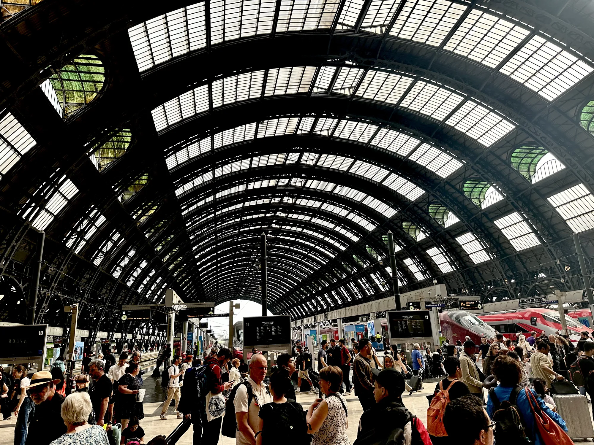 この写真はイタリアのミラノにあるミラノ中央駅で撮影されたものです。壮大なアーチ状の天井が特徴的な駅構内は、自然光が差し込む構造になっています。天井は細い鉄骨で複雑に組まれ、上部に設置された多数のガラス窓から光が差し込んでいます。駅のプラットフォームには多くの人々が行き交い、旅行者や地元の人々が混ざり合っている様子が伺えます。人々はスーツケースやバッグを持ち、それぞれの目的地に向かっているようです。ホームには電光掲示板が設置され、列車の発着情報が表示されています。背景には鮮やかな赤色の高速列車が停車しており、モダンな印象を与えています。全体的に、歴史的な建造物と現代的な交通機関が調和した、活気に満ちた駅の雰囲気が伝わってくる写真です。