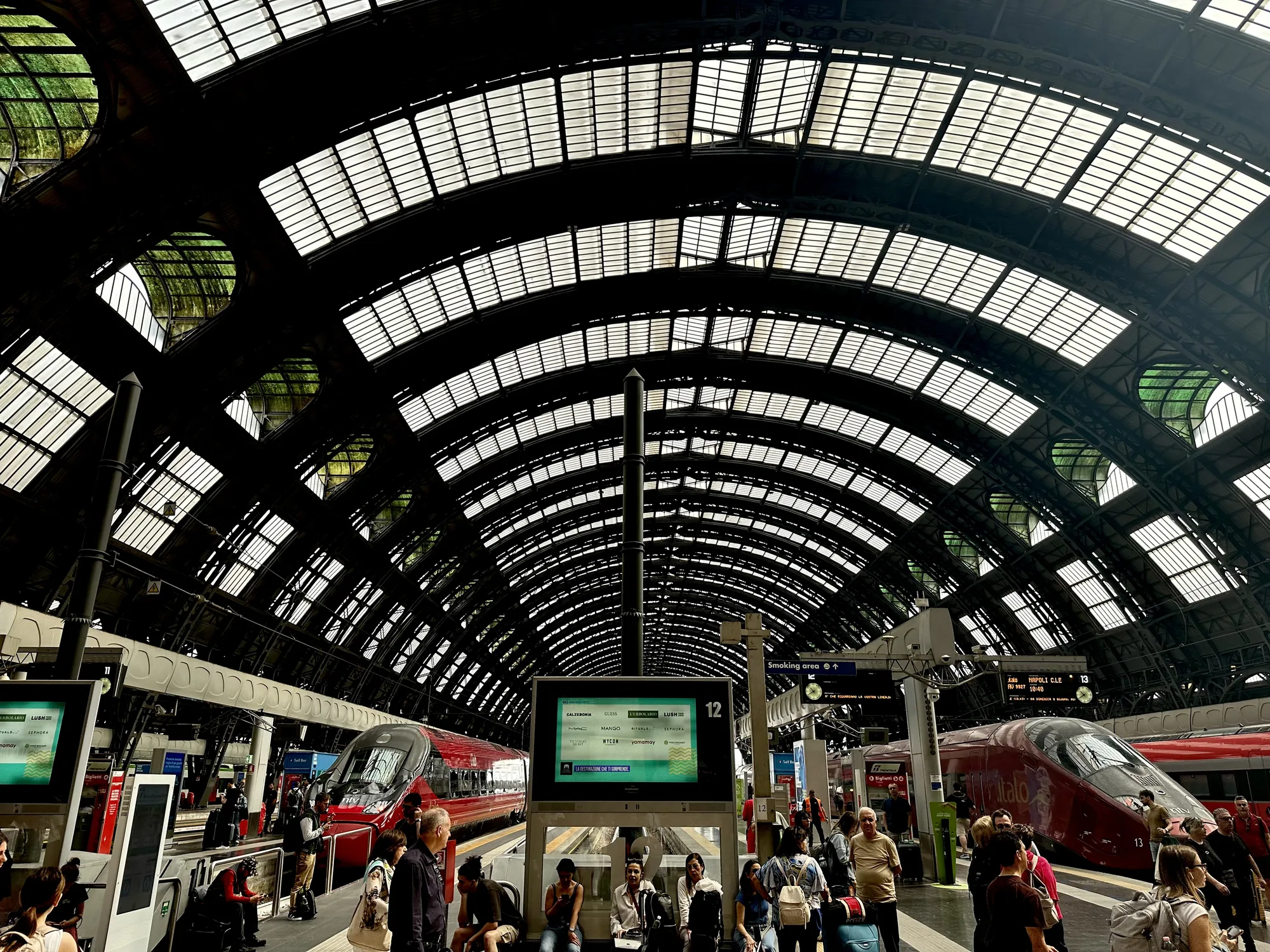 この写真はイタリアのミラノ中央駅のホームで撮影されたものです。画面中央には出発案内を表示する大きなデジタルサイネージがあり、周辺には出発を待つ人々が座ったり立ったりしています。赤色の高速鉄道Italoがホームに停車しており、多くの旅行者が列車を待っている様子が見られます。駅の天井はアーチ状の鉄骨構造で、ガラス窓がはめ込まれており、自然光が差し込んでいます。天井の複雑な構造が印象的で、歴史的な建造物であることを感じさせます。全体的に、活気があり、旅行の期待感に満ちた雰囲気です。