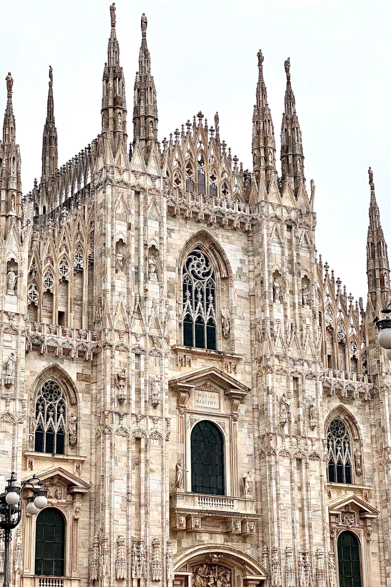 イタリアのミラノで撮影された写真には、壮大なミラノ大聖堂の一部が写っています。写真は大聖堂の正面を捉えており、その細部まで精巧なゴシック建築が際立っています。  大聖堂の壁面は、淡い色の石材で覆われており、その表面には無数の彫刻や装飾が施されています。尖塔が空に向かってそびえ立ち、その先端には彫像が配置されています。窓は大きく、美しいステンドグラスがはめ込まれているのが見て取れます。窓枠もまた、複雑な模様で飾られています。  写真全体に、重厚感と歴史的な雰囲気を感じさせます。曇り空が、建物の色調を柔らかくし、荘厳さを一層引き立てています。左下には街灯の一部が写り込んでおり、都市の一部であることを示唆しています。建築物の細部まで克明に捉えられており、その美しさに圧倒されます。