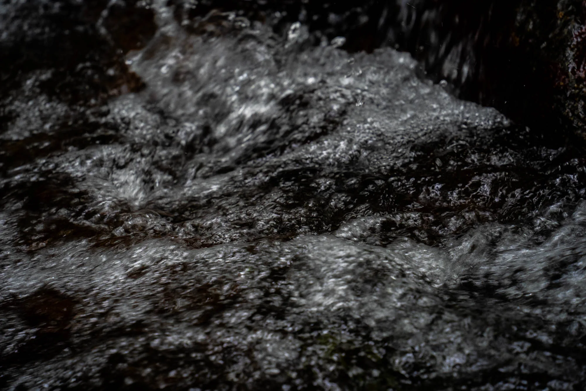 写真は、石垣島の水辺で撮影された、水流のクローズアップです。水の流れが激しく、泡立ち、表面は複雑なテクスチャを作り出しています。水の色は暗く、光の反射で部分的に白く輝いて見えます。画面右上には岩の一部が見え、それが水の流れをせき止めているように見えます。全体的に、自然の力強さと、石垣島の豊かな水を感じさせる一枚です。水の流れの速さや、水の持つ独特の質感が伝わってきます。