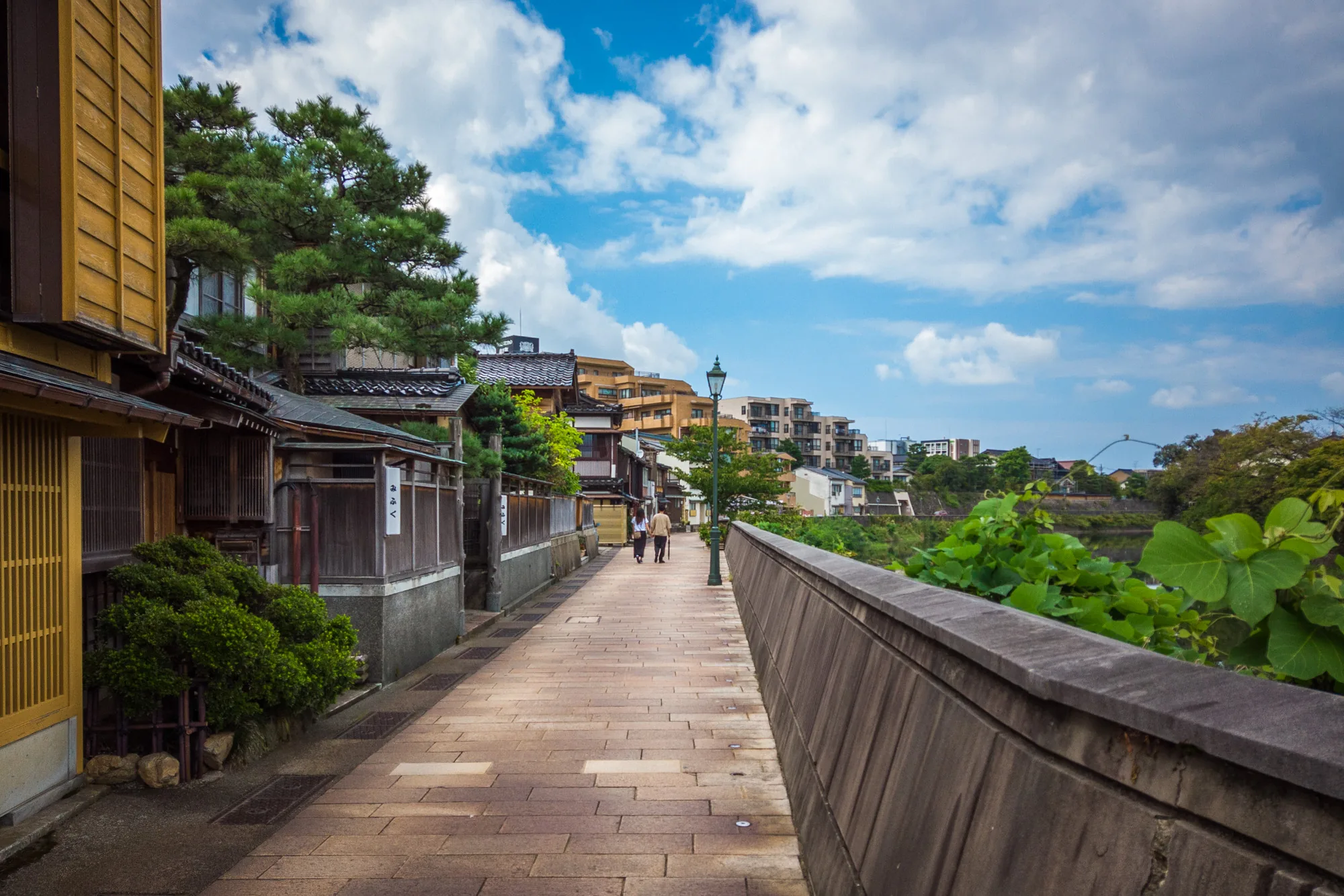 写真は日本の金沢で撮影されたもので、美しい街並みが写っています。石畳の道が奥へと伸びており、その両側には伝統的な日本家屋が建ち並んでいます。家屋の壁は木製で、一部は黄色く塗られています。屋根は瓦葺きで、緑豊かな木々が家屋を覆っています。道には緑色の街灯が立っており、遠くには現代的なマンション群が見えます。空は青く、白い雲が浮かんでいます。全体的に落ち着いた雰囲気で、歴史と現代が融合した風景です。石垣には緑の植物が生い茂り、自然の息吹も感じられます。道の奥には二人の人物が歩いており、その風景に溶け込んでいます。
