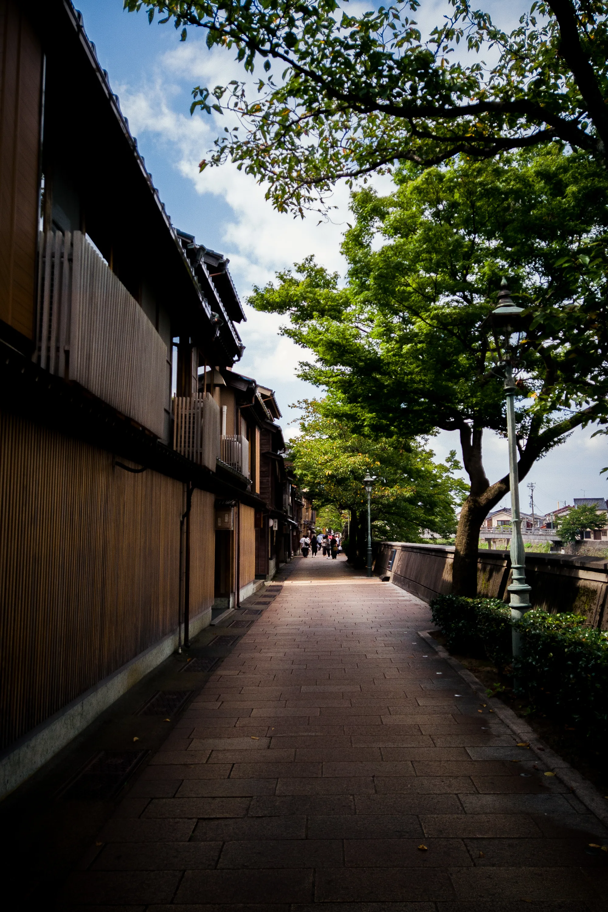 写真は石川県金沢市で撮影されたもので、趣のある古い街並みを捉えています。画面奥に向かって伸びる石畳の道は、左右を伝統的な木造家屋が囲んでいます。家屋はそれぞれ異なるデザインの格子窓や低い手すり付きのバルコニーを持ち、落ち着いた色合いの木材が使われています。  道の右側には、緑豊かな木々が生い茂り、その間には古風なデザインの街灯が立っています。遠くには、人々が歩いている様子が見え、観光地であることが伺えます。空は晴れており、一部に白い雲が見られます。全体的に、静かで落ち着いた雰囲気が感じられ、金沢の歴史と文化が色濃く残る美しい景観が表現されています。