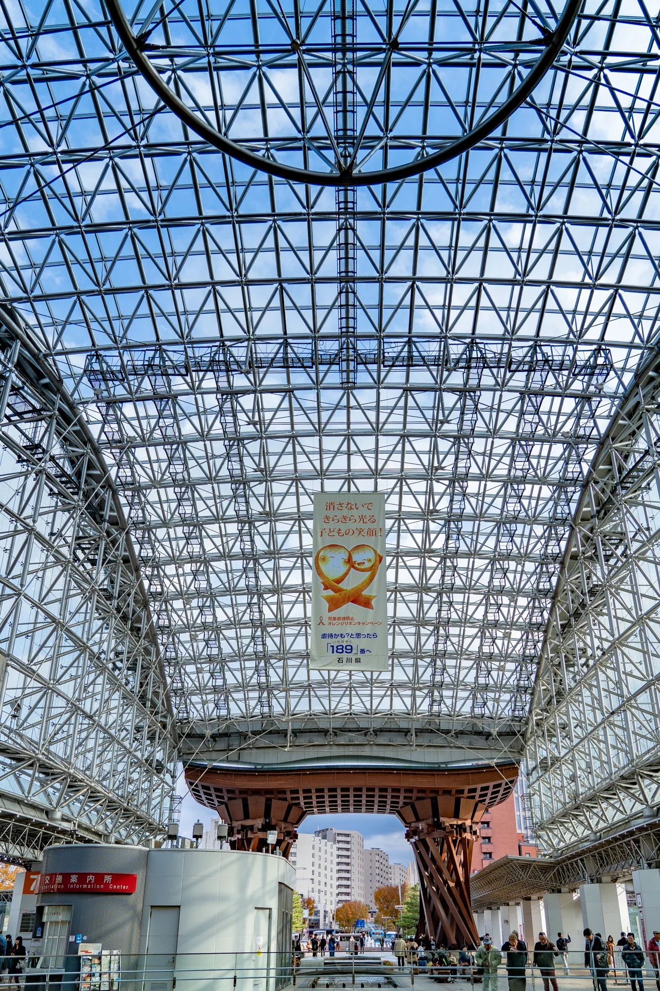この写真は、金沢駅の有名な鼓門を内側から見上げた風景を捉えています。巨大なガラスドーム状の屋根は、複雑な鋼鉄の格子構造で覆われており、その隙間から青空が覗いています。構造全体は非常に近代的で、洗練された印象を与えます。屋根の下には、木造の鼓門の一部が見えており、伝統的な要素と現代的な構造が組み合わさった独特の景観を作り出しています。また、「消さないで　きらきら光る　子どもの笑顔！」と書かれたオレンジリボンキャンペーンのバナーが吊り下げられています。これは児童虐待防止を訴えるものであり、社会的なメッセージが込められています。 写真の下部には、駅を利用する人々が歩いている様子が写っており、駅の活気ある雰囲気を伝えています。交通案内所の建物も見えます。全体的に、金沢駅のランドマークとしての鼓門の美しさと、駅の賑わいを捉えた写真と言えるでしょう。