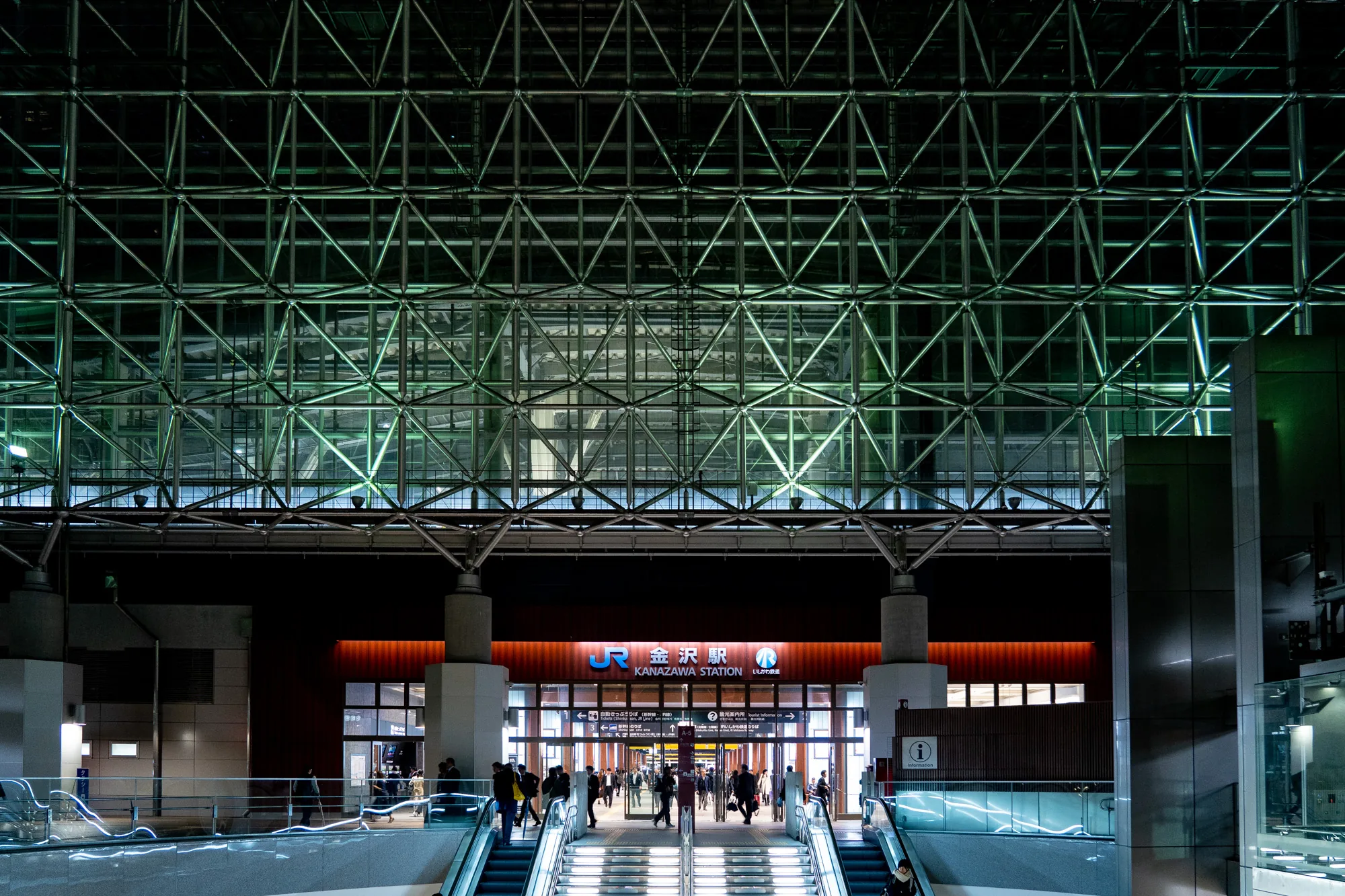 石川県金沢駅の夜の風景を捉えた写真です。印象的なのは、駅舎の天井を覆う巨大なガラスと鋼鉄の格子状の構造物で、緑色の照明によって照らされ、幾何学的な美しさを際立たせています。駅舎の入り口は、JRのロゴと「金沢駅」の文字が鮮やかに照らされ、利用客の出入りが確認できます。エスカレーターが上下に動き、人々が駅の中へと吸い込まれていく様子が写っています。駅構内は暖色の照明で照らされており、外の冷たい印象の緑色の光とのコントラストが目を引きます。全体的に、近代的で洗練された、そしてどこか幻想的な雰囲気を醸し出している写真です。