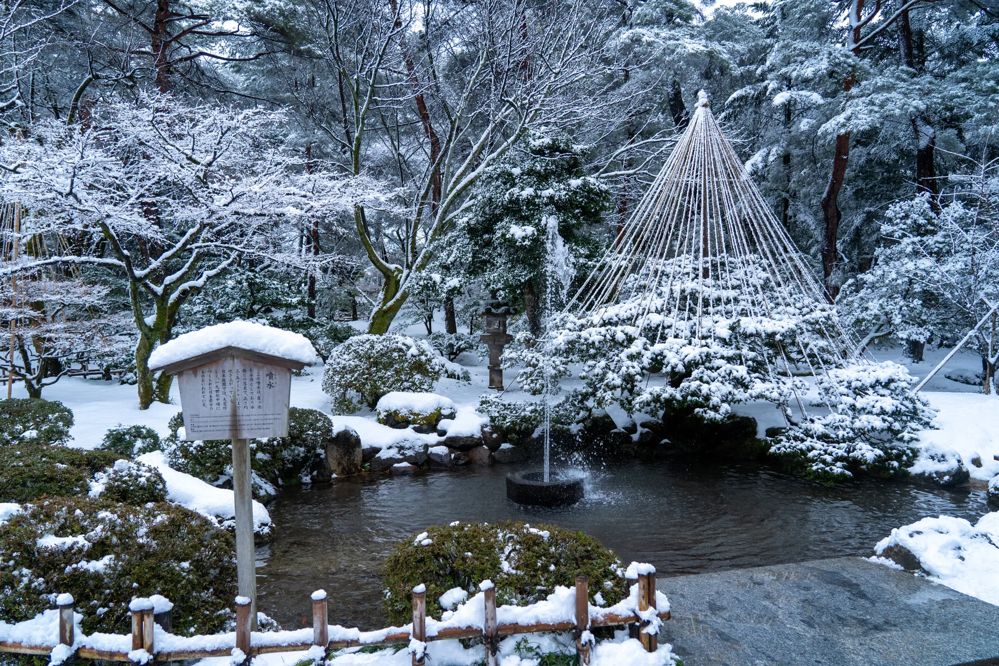 写真は、雪化粧をした日本の庭園、おそらく金沢の兼六園で撮影されたものです。庭園は静寂に包まれ、一面が雪で覆われています。池があり、中央には噴水が設置され、わずかに水面を揺らしています。噴水の周りの木々は雪の重みで枝を垂れ下げ、独特の景観を作り出しています。また、雪吊りが施された木も見られます。雪吊りは、雪の重みから木の枝を守るために施される日本庭園独特の技術です。写真には、庭園の歴史や由来を説明する案内板も写っており、雪が積もって趣を添えています。庭園の周りの木々や植物も雪に覆われ、冬の静けさを強調しています。全体的に、写真からは日本の伝統的な美しさと、冬の厳しさ、そして静謐さが伝わってきます。OCRの結果から噴水の説明文が読み取れます。この噴水は霞ヶ池を水源としており、高さ約3.5メートルまで吹き上がるとのこと。日本庭園では珍しい19世紀中頃に作られた日本最古のものと書かれています。