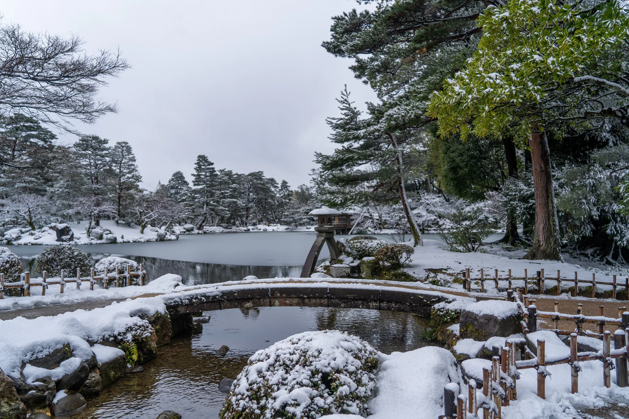 写真は、日本の石川県金沢市にある兼六園の雪景色を捉えています。空は厚い雲に覆われ、雪が降った直後のようです。庭園全体が白く覆われており、静寂で幻想的な雰囲気が漂っています。手前には、雪化粧をした丸みを帯びた植え込みがあり、その奥には石橋が架かっています。橋の下には清らかな水が流れ、雪解け水が流れ込む様子がうかがえます。  池の一部は凍り付いており、水面に雪が薄く積もっています。池のほとりには、雪をまとった木々が立ち並び、背景には針葉樹林が広がっています。池の中央には、雪に覆われた灯籠が佇み、庭園の景観に趣を添えています。写真全体から、冬の兼六園の静けさと美しさが伝わってきます。雪の白さと木々の緑のコントラストが印象的です。