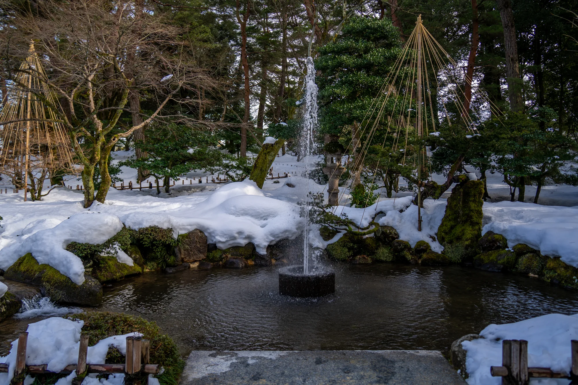 金沢の兼六園と思われる雪景色を捉えた写真です。中央には噴水のある池があり、水面は静かで落ち着いた色合いをしています。噴水は勢いよく水を噴き上げており、その水しぶきが周囲の空気を潤しているようです。  背景には、雪を被った庭園の木々が並んでいます。雪吊りが施された木々もあり、冬の寒さから枝を守るための工夫が見られます。地面は一面雪に覆われており、緑色の苔むした岩とのコントラストが美しいです。  全体的に静寂で落ち着いた雰囲気で、雪の白さが一層その雰囲気を際立たせています。日本の冬の美しさを感じさせる一枚です。