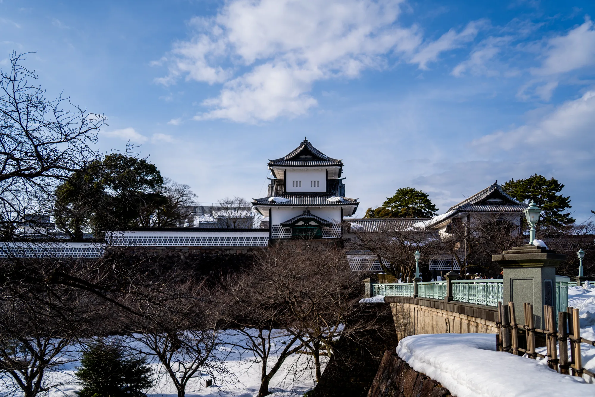 写真は、日本の金沢で撮影された金沢城の冬の風景です。中央には白壁と黒い屋根が特徴的な城の建物が堂々とそびえ立っています。屋根にはうっすらと雪が積もり、冬の寒さを感じさせます。建物の周囲には、白と黒の模様が施された塀が巡らされており、歴史的な雰囲気を醸し出しています。手前には雪が積もった川と、枝を広げた冬の木々が写っており、モノクロームに近い色合いが静寂さを強調しています。空は晴れており、白い雲が浮かんでいます。全体的に、日本の歴史と自然が調和した美しい風景写真です。