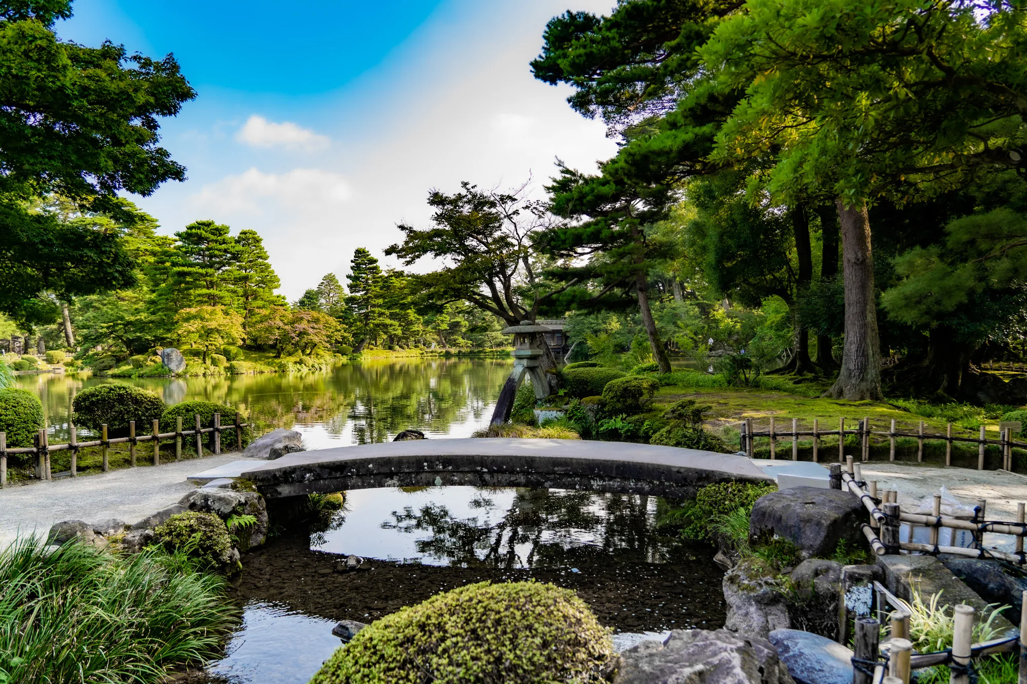 写真は、日本の金沢にある庭園で撮影されたものです。画面中央には、苔むした岩でできた小さな橋が架かっており、その下を清らかな水が流れています。橋の奥には池が広がり、周囲には緑豊かな木々が生い茂っています。空は青く、白い雲が浮かんでおり、穏やかな日差しが庭園全体を照らしています。橋のたもとには、丸く刈り込まれた植え込みや石が配置され、繊細な景観を作り出しています。全体的に落ち着いた雰囲気で、日本の伝統的な美しさを感じさせる風景です。遠景には灯篭も見え、庭園の趣を深めています。