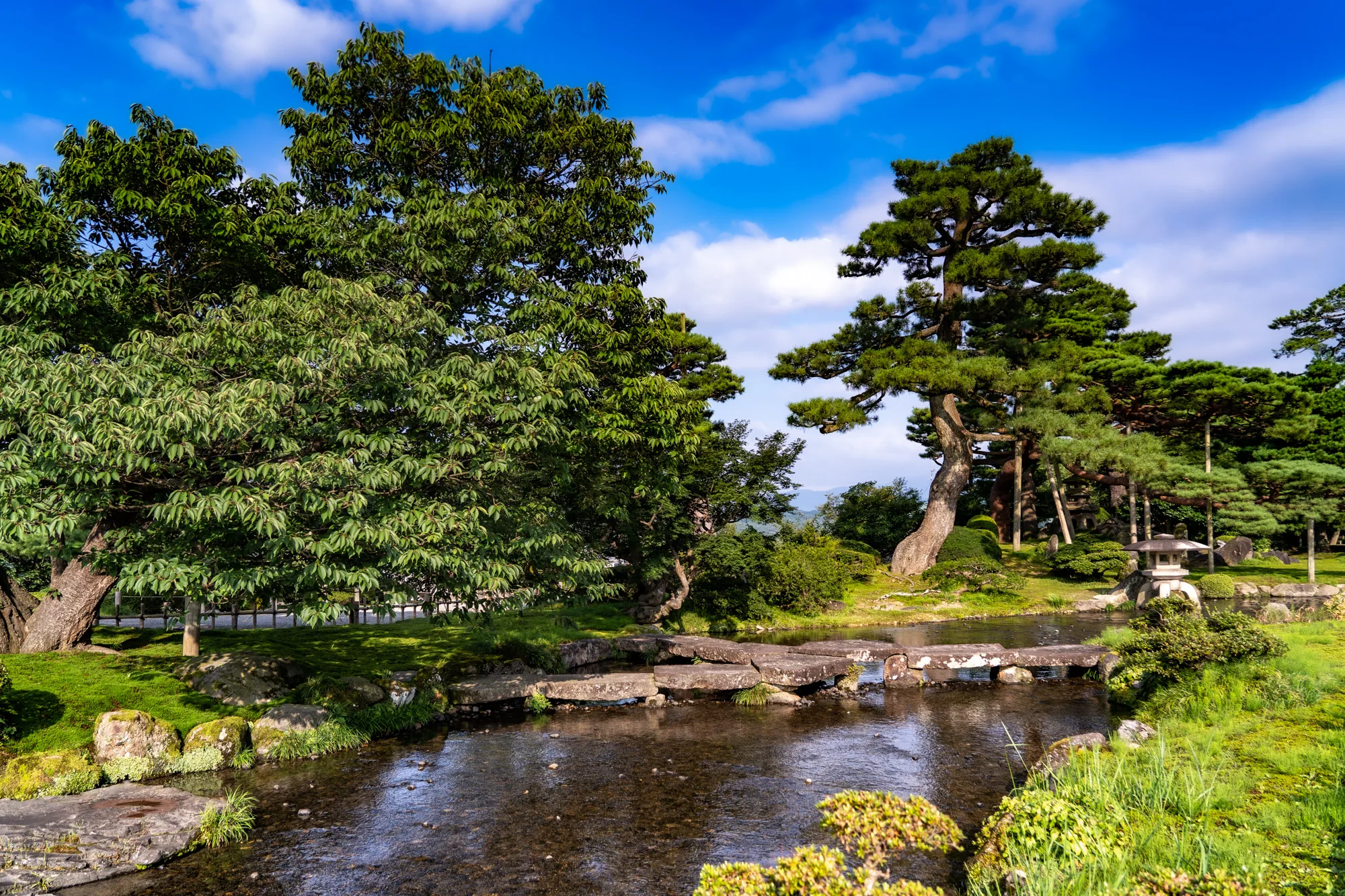 写真は日本の金沢で撮影された庭園の風景を捉えています。澄み切った青空と白い雲が広がり、豊かな緑が目に鮮やかです。前景には、苔むした石でできた橋が小さな川を渡っており、水面は光を反射してキラキラと輝いています。川の両岸には緑豊かな草木が生い茂り、石灯籠が静かに佇んでいます。背景には、堂々とした松の木や様々な種類の木々が茂り、奥行きのある景色を作り出しています。全体的に、穏やかで静寂な日本の庭園の雰囲気が伝わってくる写真です。手入れの行き届いた庭園の美しさが、見る人の心を癒してくれるような印象を与えます。