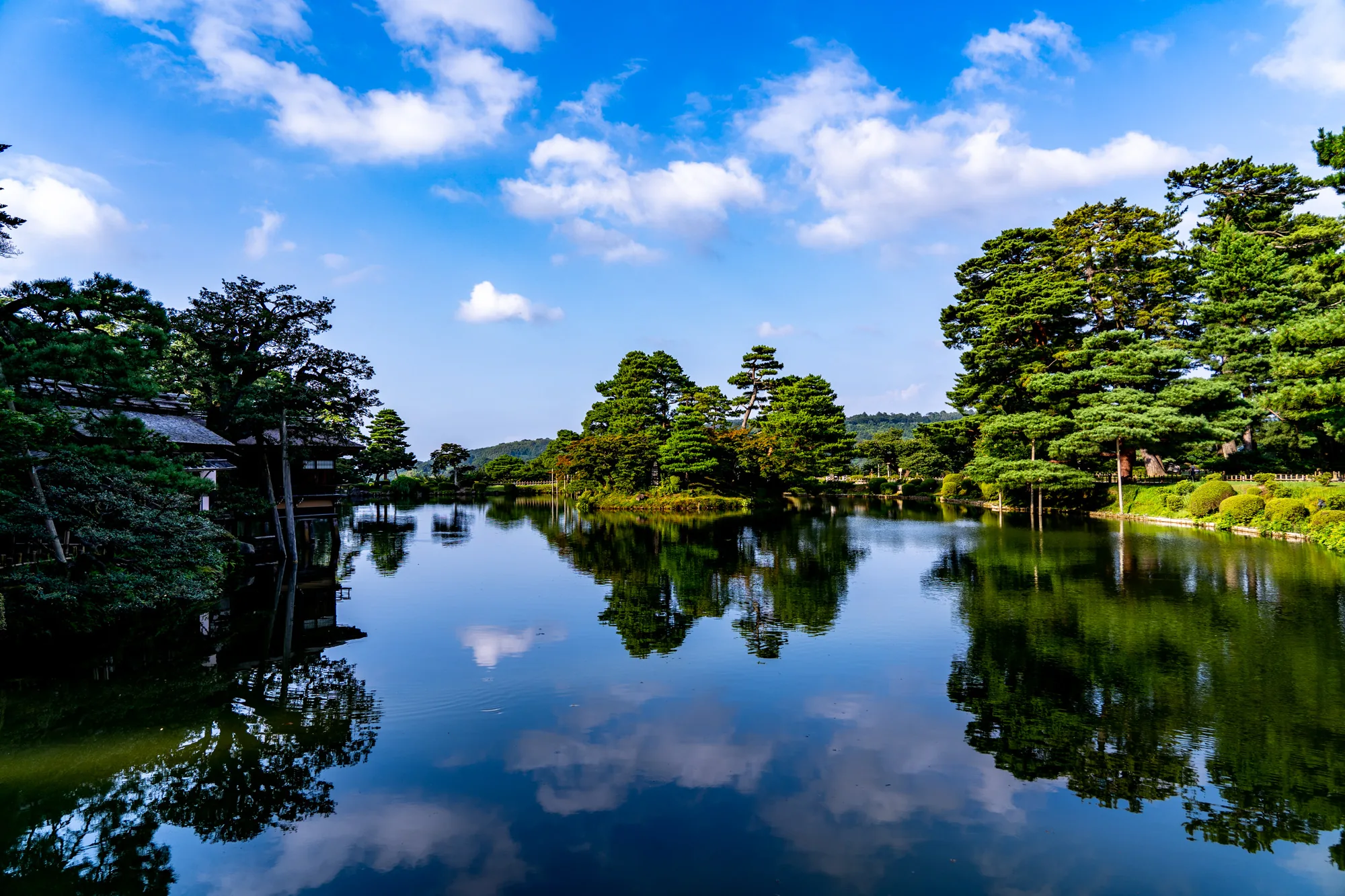 写真は日本の金沢で撮影された庭園の風景を捉えています。中心には穏やかな水面を持つ大きな池があり、空と周囲の景色を鏡のように反射しています。空は晴れ渡り、白い雲が点在しており、その姿が水面に映り込んでいます。池の周りには、手入れの行き届いた緑豊かな木々が茂っており、様々な形や大きさの木々が庭園の景観を豊かにしています。池の端には、伝統的な日本の建物がわずかに見え、その独特な屋根が特徴的です。写真全体から、静かで落ち着いた雰囲気が伝わってきます。水面に映る景色が美しく、日本の伝統的な美意識を感じさせる一枚です。